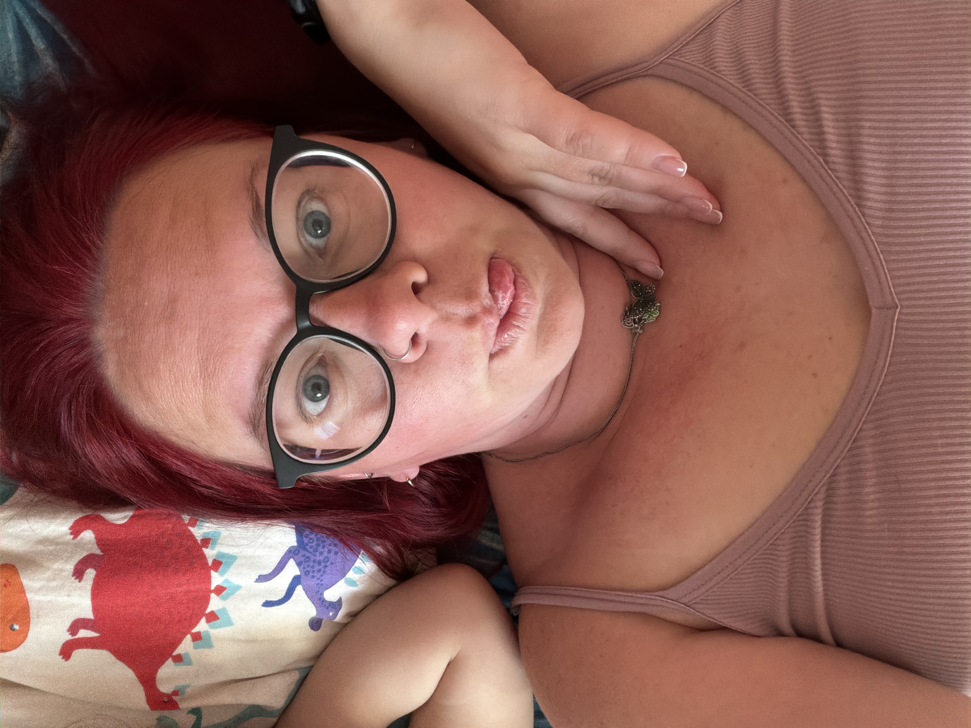 A woman with red hair is lying on a bed. She is wearing black eyeglasses, a light pink ribbed tank top, and a necklace with a small pendant. Her right hand is near her neck. The bed has a pillow with a dinosaur pattern. The city location is not discernible from the image.