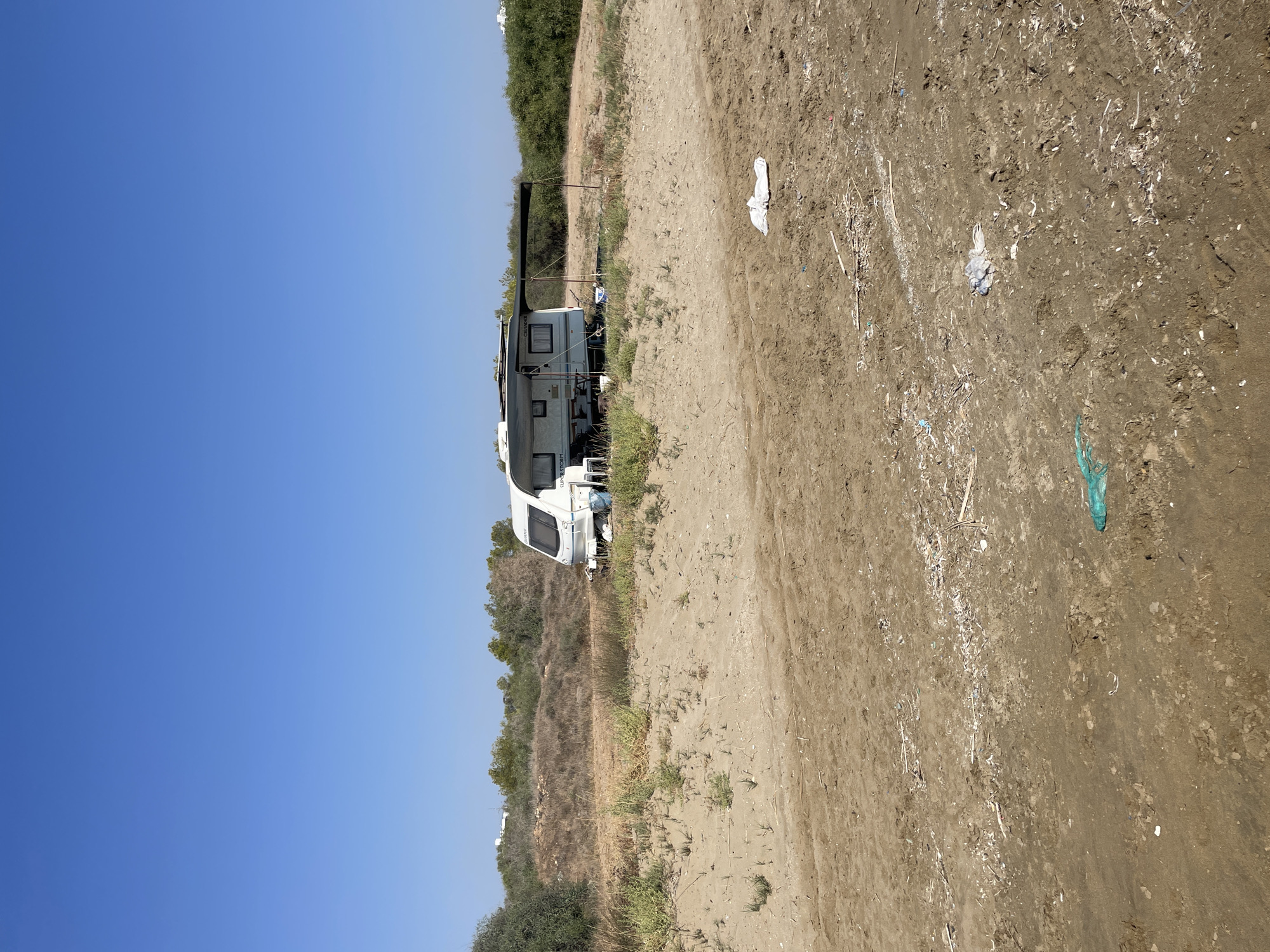 A white caravan is parked on a sandy area with some vegetation. The caravan has an awning extended and some chairs are set up outside. In the foreground, the sandy ground is littered with debris including plastic bags and what appears to be broken shells and sticks. The sky is clear and blue. The location is Spathariko, Northern Cyprus / Cyprus.
