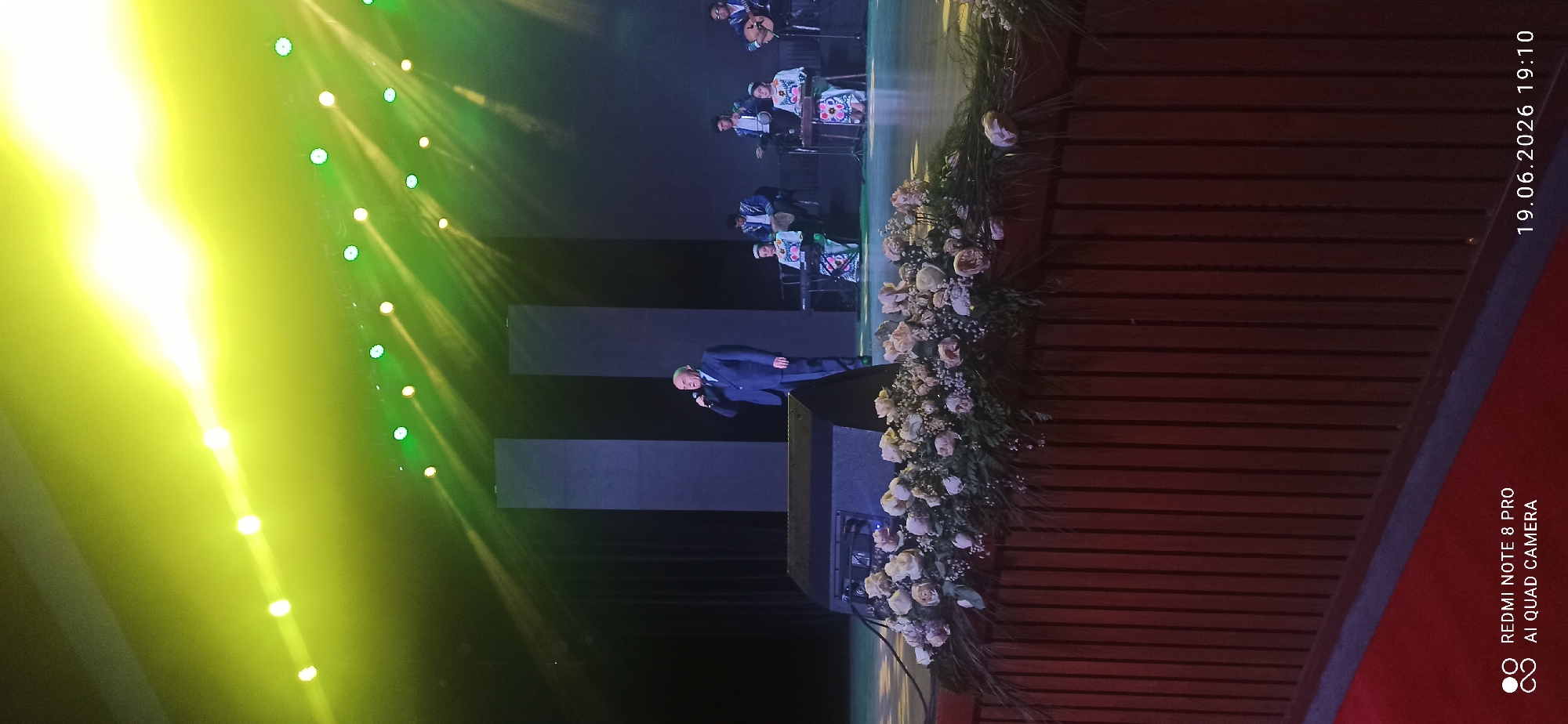 A concert stage is set with a male singer in a dark suit holding a microphone, standing center stage. Behind him, several musicians are seated, some playing instruments like a dulcimer and a string instrument, while others are seated at keyboards. The stage is illuminated by bright green and yellow lights, casting beams across the dark backdrop. In front of the stage, a wooden podium is adorned with a large floral arrangement of roses and smaller flowers, and a speaker is visible on the left side. A red carpet runs along the bottom of the image. The date and time "19.06.2026 19:10" and "REDMI NOTE 8 PRO AI QUAD CAMERA" are watermarked in the bottom right and left corners, respectively.