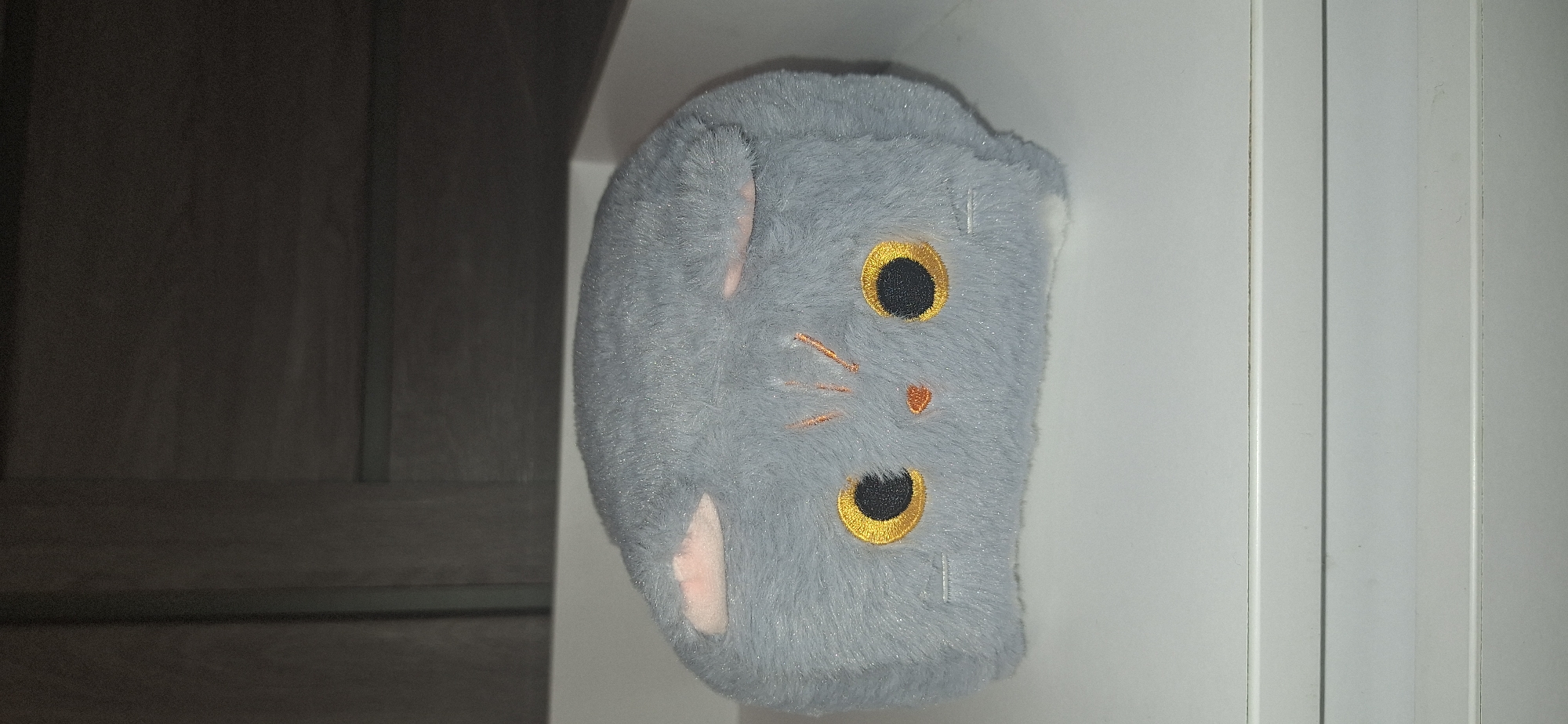 A plush toy resembling a gray cat is positioned on a white surface. The toy has large, round yellow eyes with black pupils and embroidered orange whiskers and a nose. Its ears are light pink. The background is a dark gray, textured surface, possibly a wall or cabinet. The lighting suggests the photo was taken indoors. The location is not specified in the image.