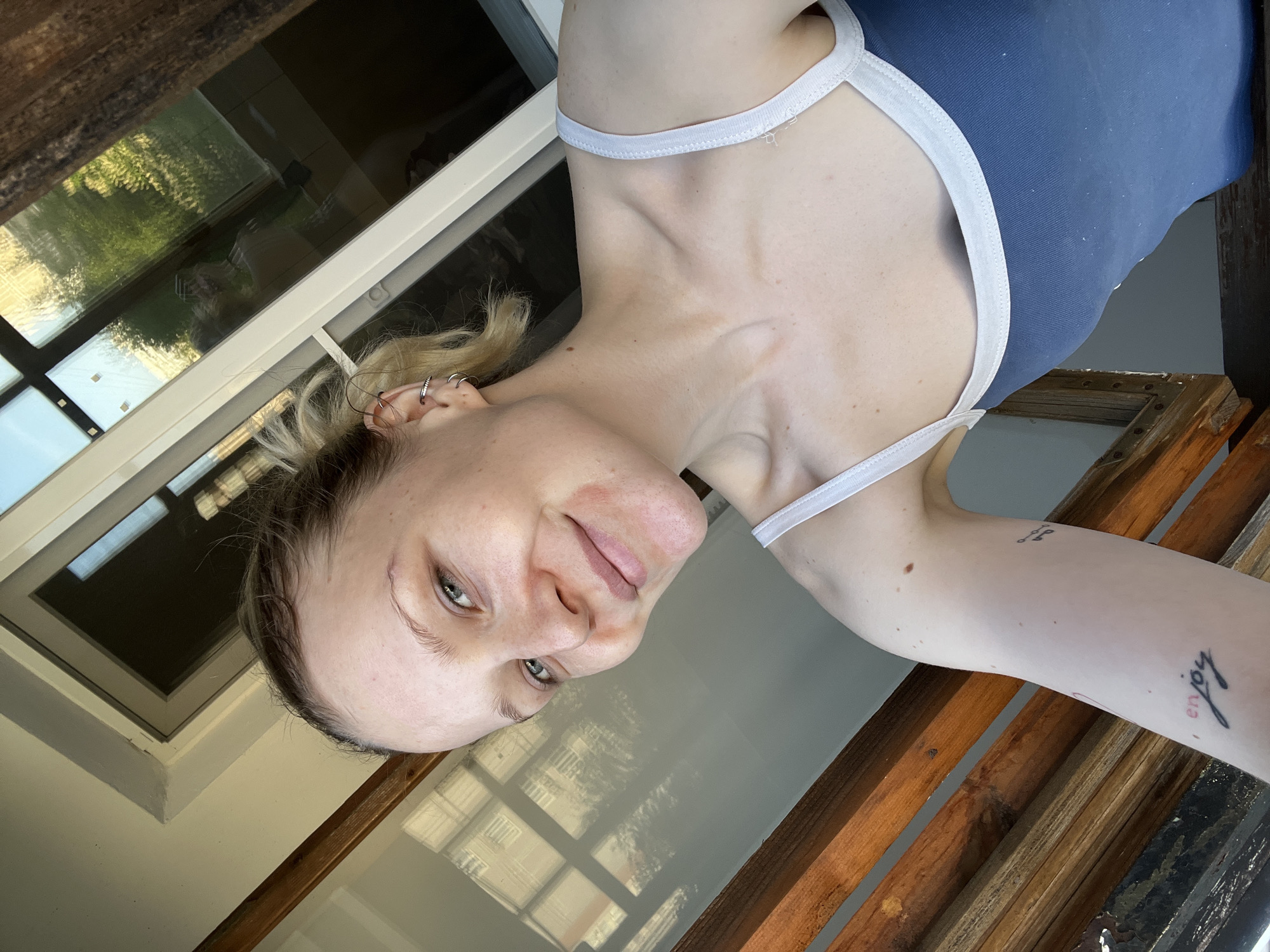 A light-skinned person with blonde hair and multiple ear piercings is the primary subject of this selfie. They are wearing a blue tank top with white trim. The person appears to be standing either indoors or on a porch as indicated by the wooden railing to their left. Through a window behind the person, trees can be seen outside. The person has tattoos on their left arm. The location is not discernable from the image and therefore cannot be said to be located in Khimki, Russia.