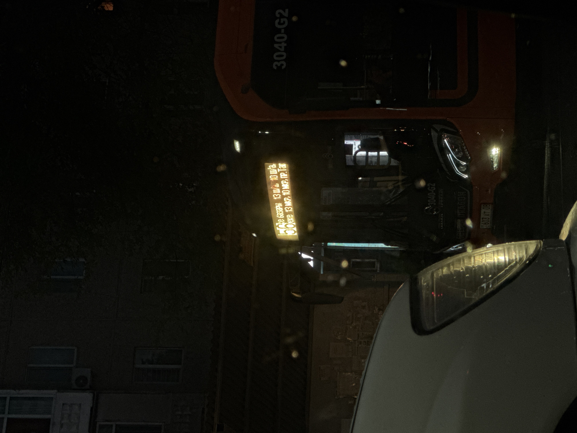 The image captures a nighttime scene in the city of Taraz, Kazakhstan, near the city administration building. In the foreground, the headlight of a light-colored vehicle is visible on the left side. To the right, an orange bus is prominently featured, displaying the number "3040-G2" on its front. A yellow illuminated sign on the bus displays Cyrillic text, likely indicating route information or destination. The background shows a multi-story building with windows and an air conditioning unit, partially obscured by trees and darkness. The atmosphere is illuminated by artificial lights from the bus and the building, with some light reflections and water droplets visible on the vehicle's surfaces. The overall impression is of a street scene at night, with vehicular traffic and urban infrastructure.