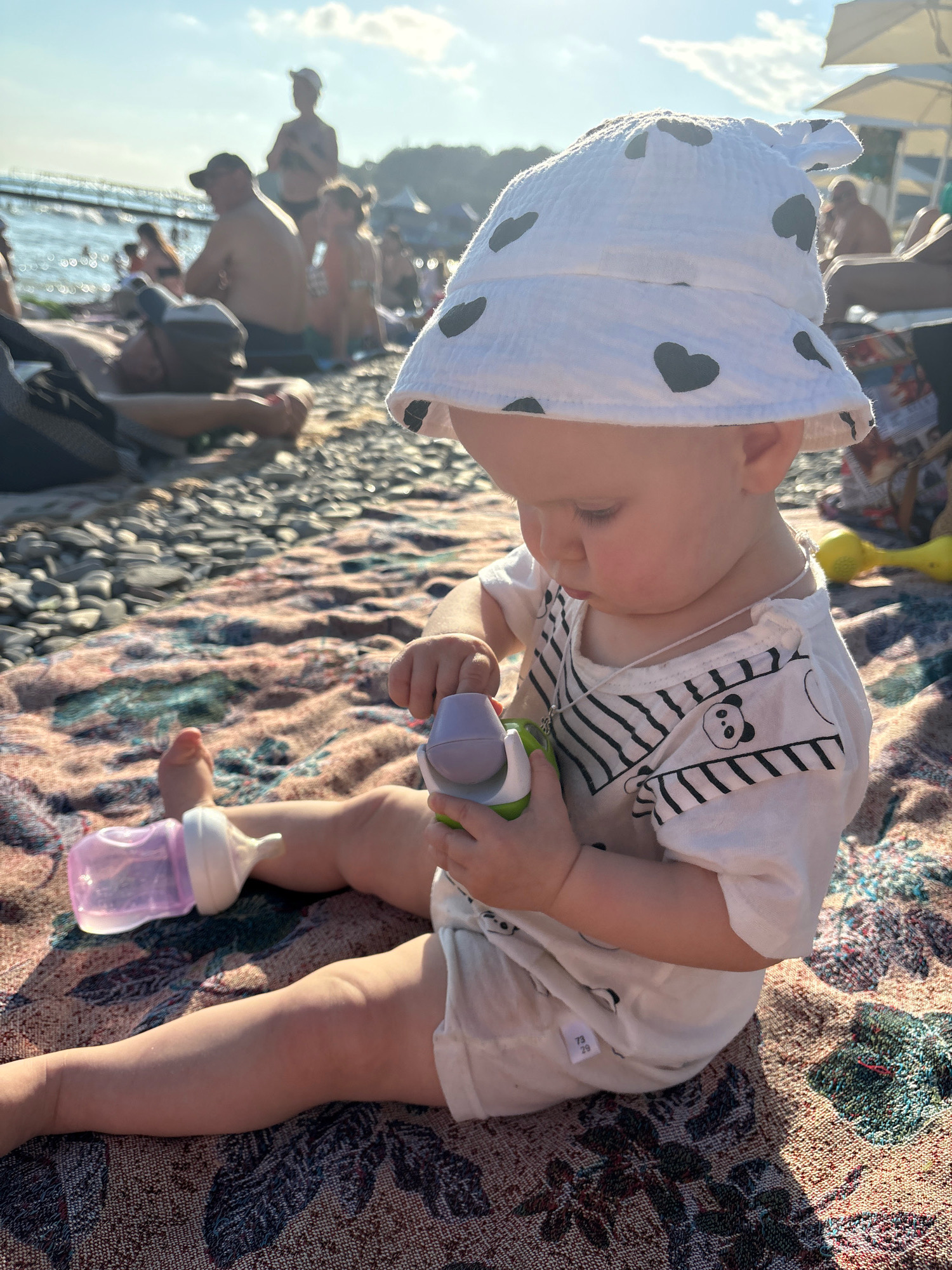 A baby is sitting on a patterned blanket on a pebble beach in Arkhipo-Osipovka, Russia. The baby is wearing a white bucket hat with black hearts and a white t-shirt with black stripes and a panda graphic. The baby is holding a purple and green toy. A pink baby bottle with a white nipple is lying on the blanket near the baby's left leg. In the background, other people are sitting and lying on the beach, and the sea is visible. The sun is shining, casting a warm glow on the scene.