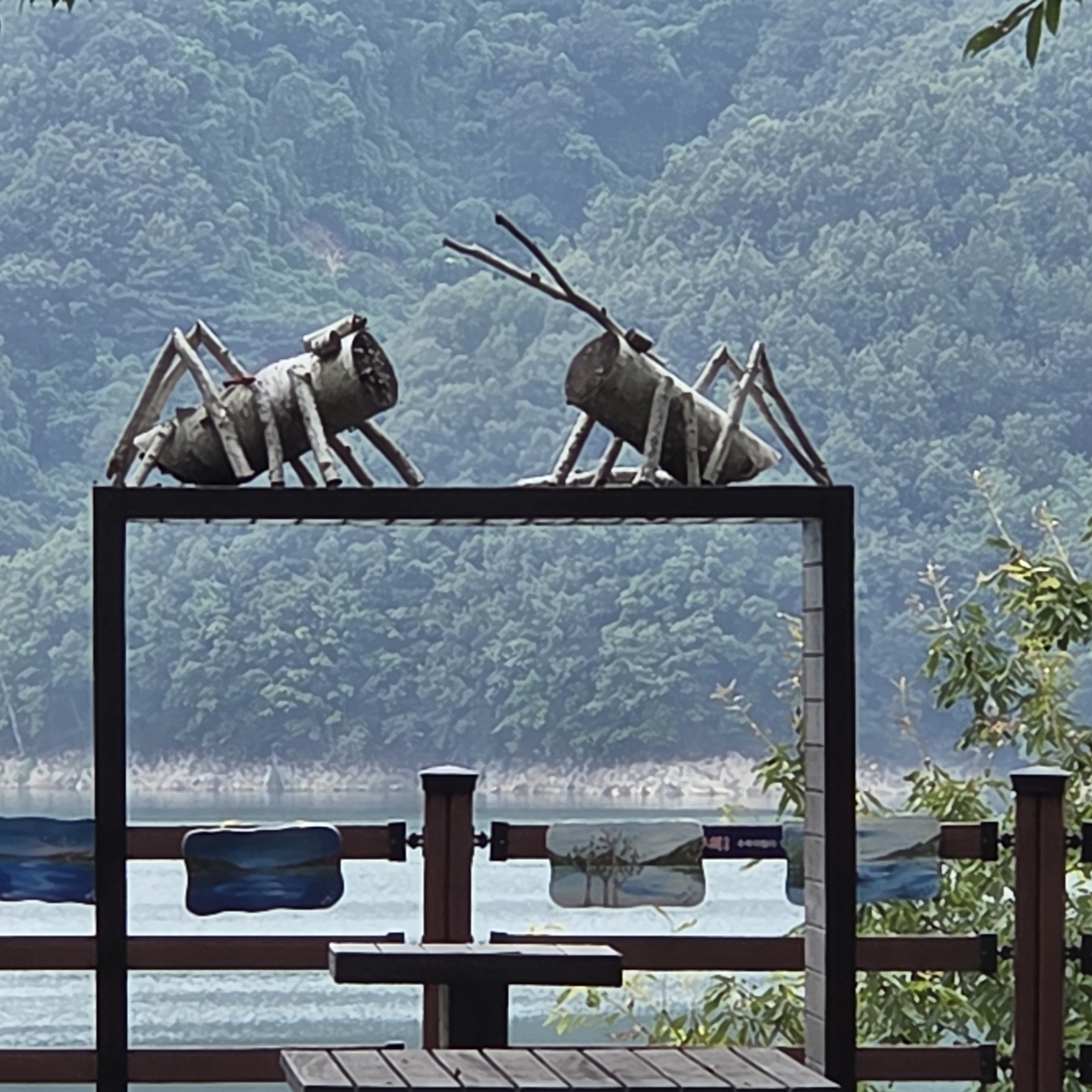 The image depicts an outdoor scene featuring two large, stylized insect sculptures made from logs, positioned on a dark metal platform. The sculptures are situated in front of a body of water and a densely wooded hillside. In the foreground, a wooden railing with decorative plaques depicting landscapes separates the viewer from the water. A wooden picnic table is also visible in the foreground. The overall impression is of an artistic installation in a natural setting. The location is identified as Gapcheon-myeon, South Korea. There are no people visible in the image.