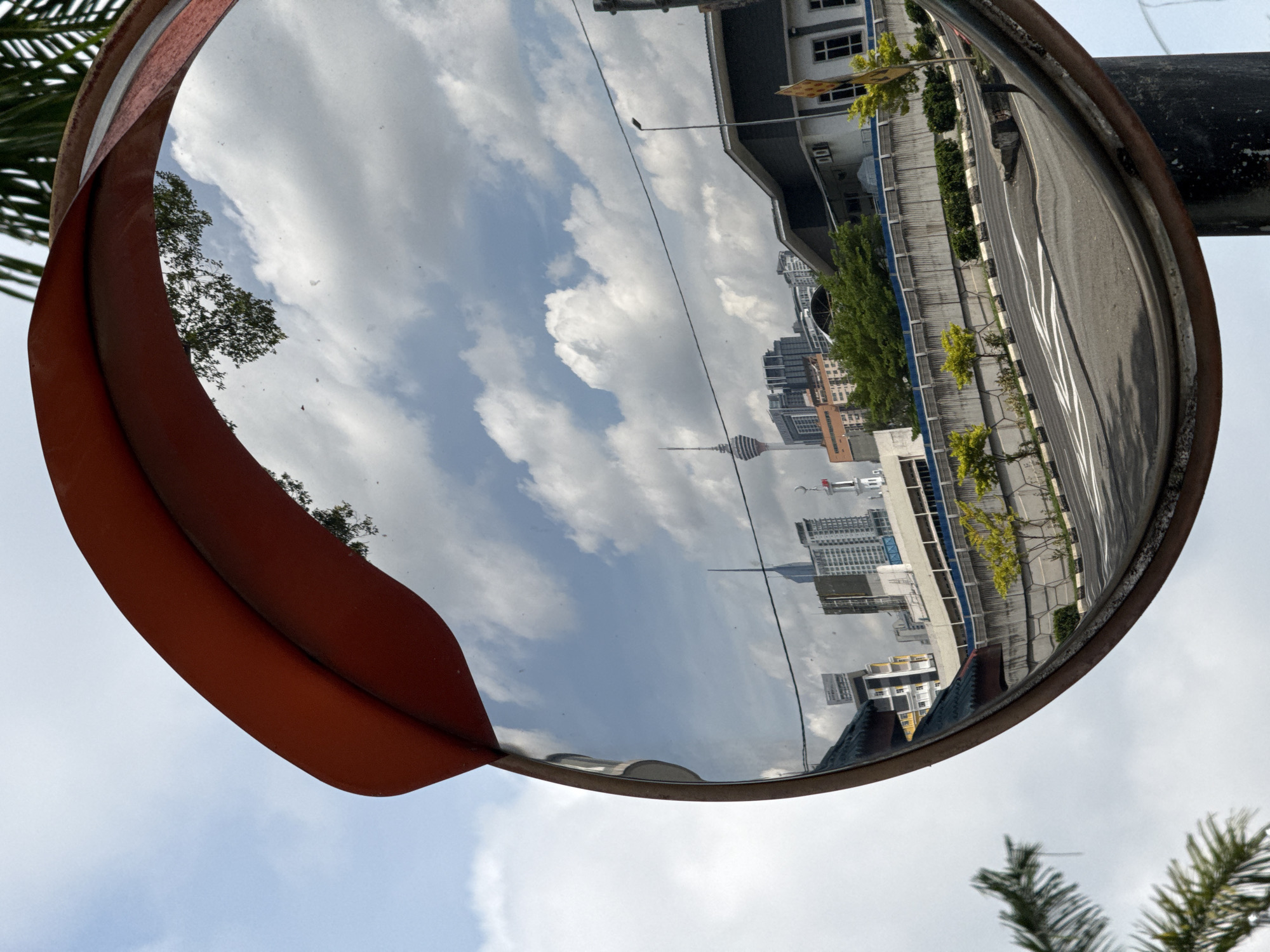 The image shows a convex traffic mirror reflecting a cityscape. The Kuala Lumpur Tower (Menara Kuala Lumpur) is visible among other buildings. A road with painted lines and a sidewalk with trees runs alongside buildings. The sky is visible with scattered clouds. The mirror is mounted on a pole. The mirror has an orange visor. Palm tree branches are visible in the foreground.