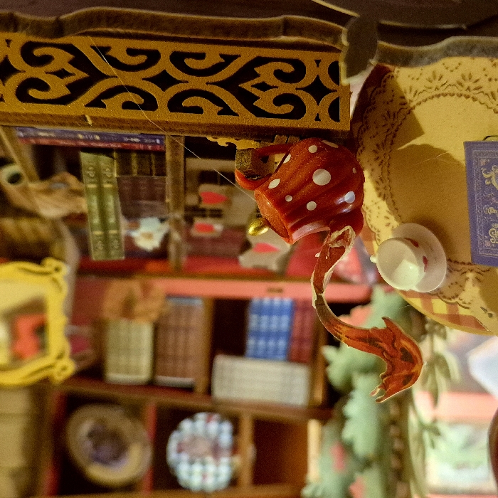 The image displays a close-up view of a miniature scene, likely a dollhouse or diorama, characterized by whimsical and fantastical elements, reminiscent of Alice in Wonderland. The perspective is angled, focusing on the upper and mid-sections of the miniature setup.
In the upper part of the frame, there is a horizontally oriented, golden-brown wooden structure with an intricate, dark-colored filigree pattern cut into it, resembling a decorative frieze or shelf border. A thin, light-colored string or thread is visible, stretching across this patterned structure.
Hanging upside down from the golden-brown structure is a vibrant red teacup adorned with white polka dots, giving it the appearance of a mushroom cap. From the rim of this teacup, a translucent, fiery orange and yellow element extends downwards in a wavy, fluid shape, suggesting a pouring liquid or a magical stream.
To the right of the red teacup, a circular, cream-colored surface with an embossed or patterned texture, resembling a doily or tablecloth, is visible. Perched on this surface, also appearing to be somewhat suspended or precariously placed, is a tiny white teacup. Next to it, a small, rectangular object with a dark blue background and intricate gold-colored patterns is present, possibly a miniature book or decorative plaque.
The background is filled with miniature furniture, primarily bookshelves, which are reddish-brown. These shelves are packed with numerous miniature books of various colors, including green, blue, and brown, with some spines faintly showing details. Other small, decorative items are also visible on the shelves, such as what appear to be miniature vases, bowls, and a small plate with a distinctive patterned design. On the far left, a small, out-of-focus golden picture frame is visible. Towards the lower right, out-of-focus green elements suggest miniature foliage or plants.
The lighting appears warm and somewhat diffused, enhancing the intimate and detailed nature of the miniature display. The overall impression is one of an enchanted or magical setting, rich in small details that invite closer inspection. There is no visible text within the image.
