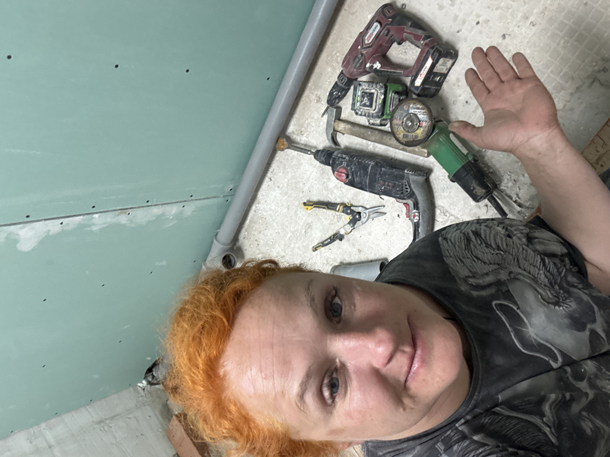 The image shows a person with orange hair, fair skin, and blue eyes, wearing a dark gray patterned shirt. They are in a room with pale green drywall on the wall. Various tools are scattered on the floor, including a hammer drill, angle grinder, laser level, hammer, and wire cutters. There is a gray pipe running along the wall. The location is unspecified, but the prompt suggests it is Chaikovsky, Russia. The person is looking at the camera.
