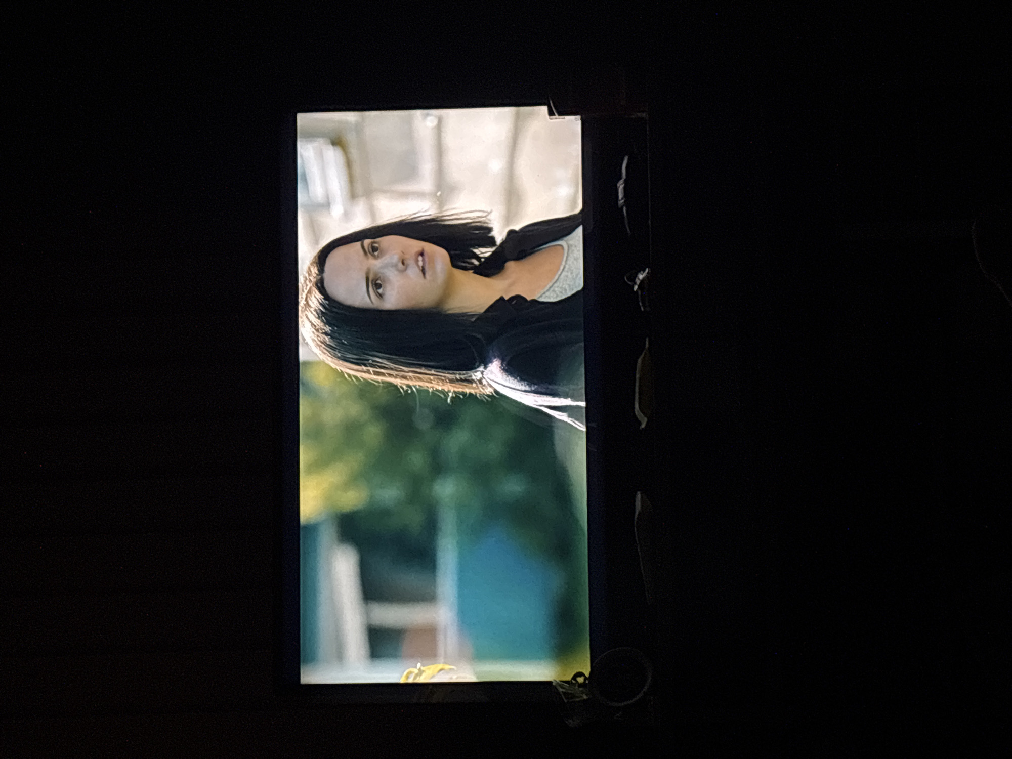 The image shows a television screen displaying a scene from a movie or show. The scene features a young woman with dark, straight hair and a serious expression, looking off to the right. She is wearing a dark jacket over a light-colored shirt. The background behind her is blurred, suggesting an outdoor setting with buildings and greenery. The television itself is positioned in a dark room, with only the illuminated screen and a portion of the bottom of the TV visible. A small, circular object is attached to the left side of the television bezel. The overall impression is of someone watching content on a screen in a dimly lit environment.