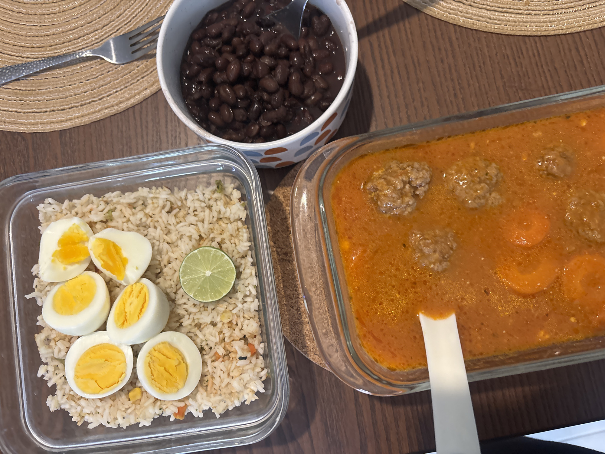 A tabletop view presents a meal comprised of rice with halved boiled eggs and a lime wedge in a clear glass container, a bowl of black beans with a fork, and a separate clear glass container of soup with meatballs and sliced carrots. These items are placed on a wooden table with a circular woven placemat visible. There is no clear indication of the location in Puebla, Mexico, as no distinct geographical landmarks or cultural references are present.