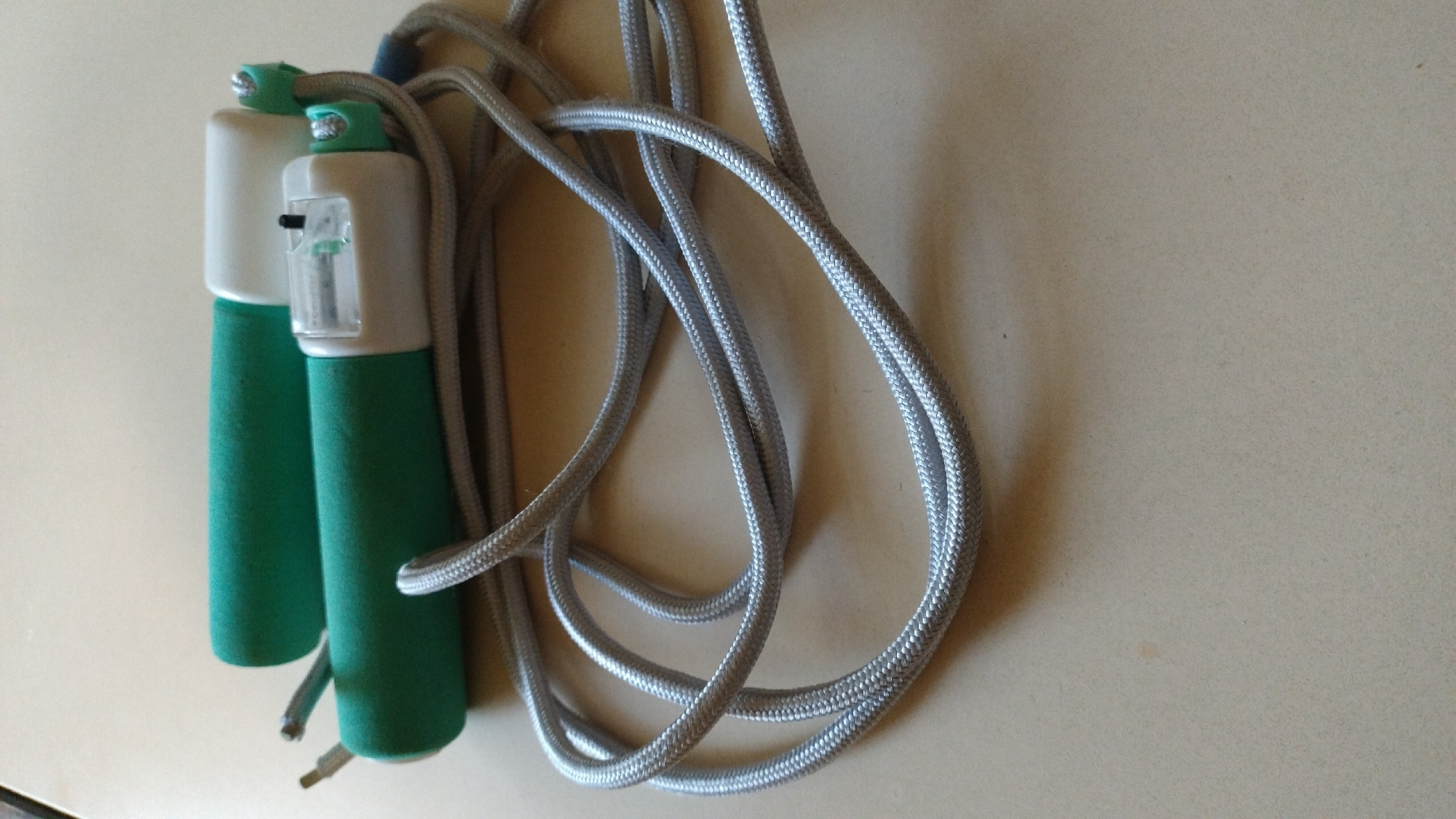A jump rope with green foam handles and a silver rope lies on a light brown, textured surface. One handle has a clear plastic counter with a black button. The rope is coiled loosely around the handles, with the ends of the rope extending out from the left side of the handles. The background is out of focus. The scene is indoors, with no indication of location in a city.
