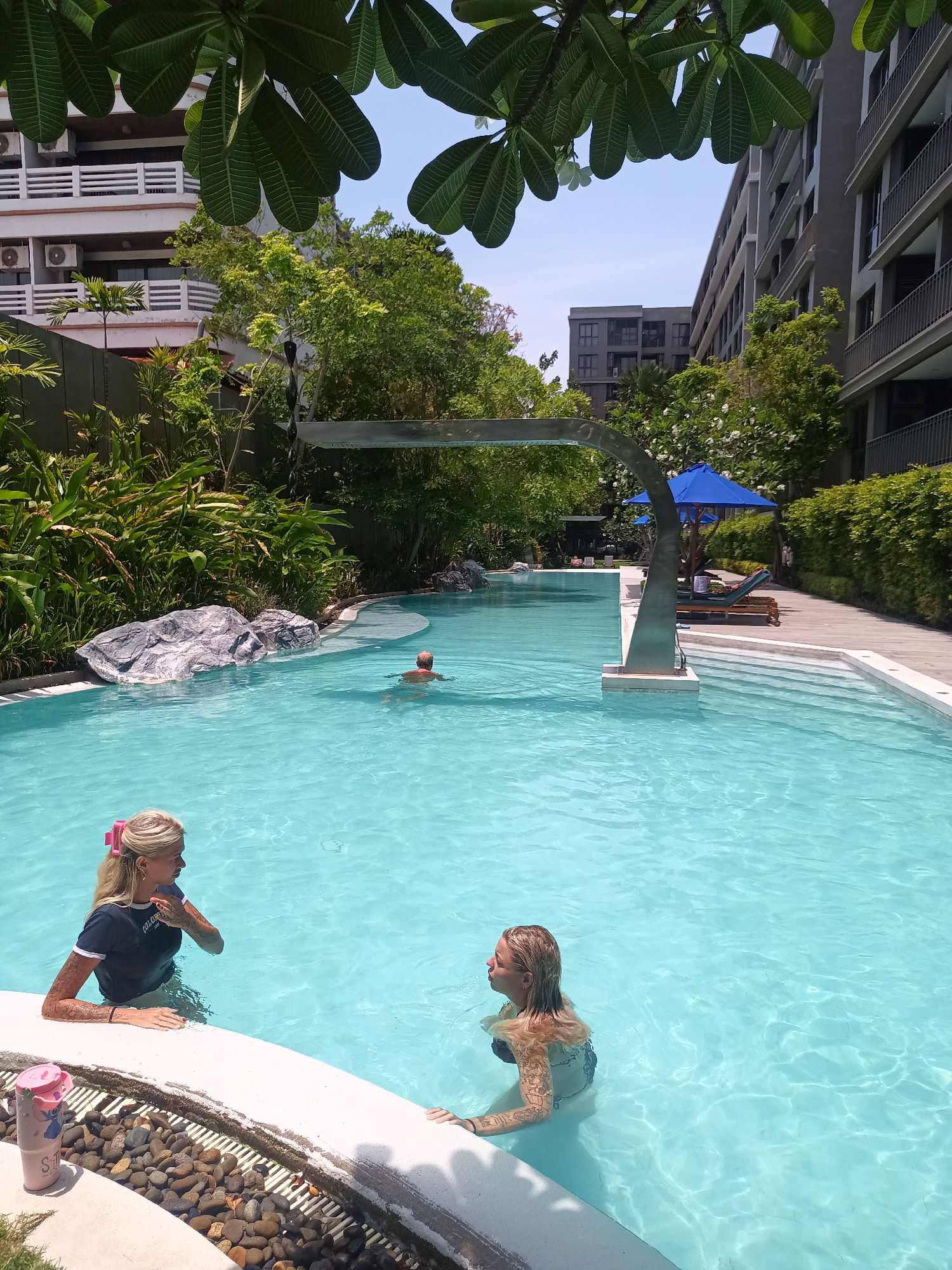 The image shows a swimming pool at a resort in Hua Hin City Municipality, Thailand. Two women are in the foreground; one with blonde hair and a pink clip, wearing a dark-colored shirt, is standing in the shallow part of the pool near the edge. The other woman, also blonde, is swimming in the pool with her arms and shoulder showing, facing right. A man is swimming in the background. There are trees and plants surrounding the pool, and buildings can be seen in the background. Lounge chairs with blue umbrellas are visible to the right of the pool. A silver arched structure is over the pool in the mid-ground. A child's pink drink container is sitting on the ledge of the pool.