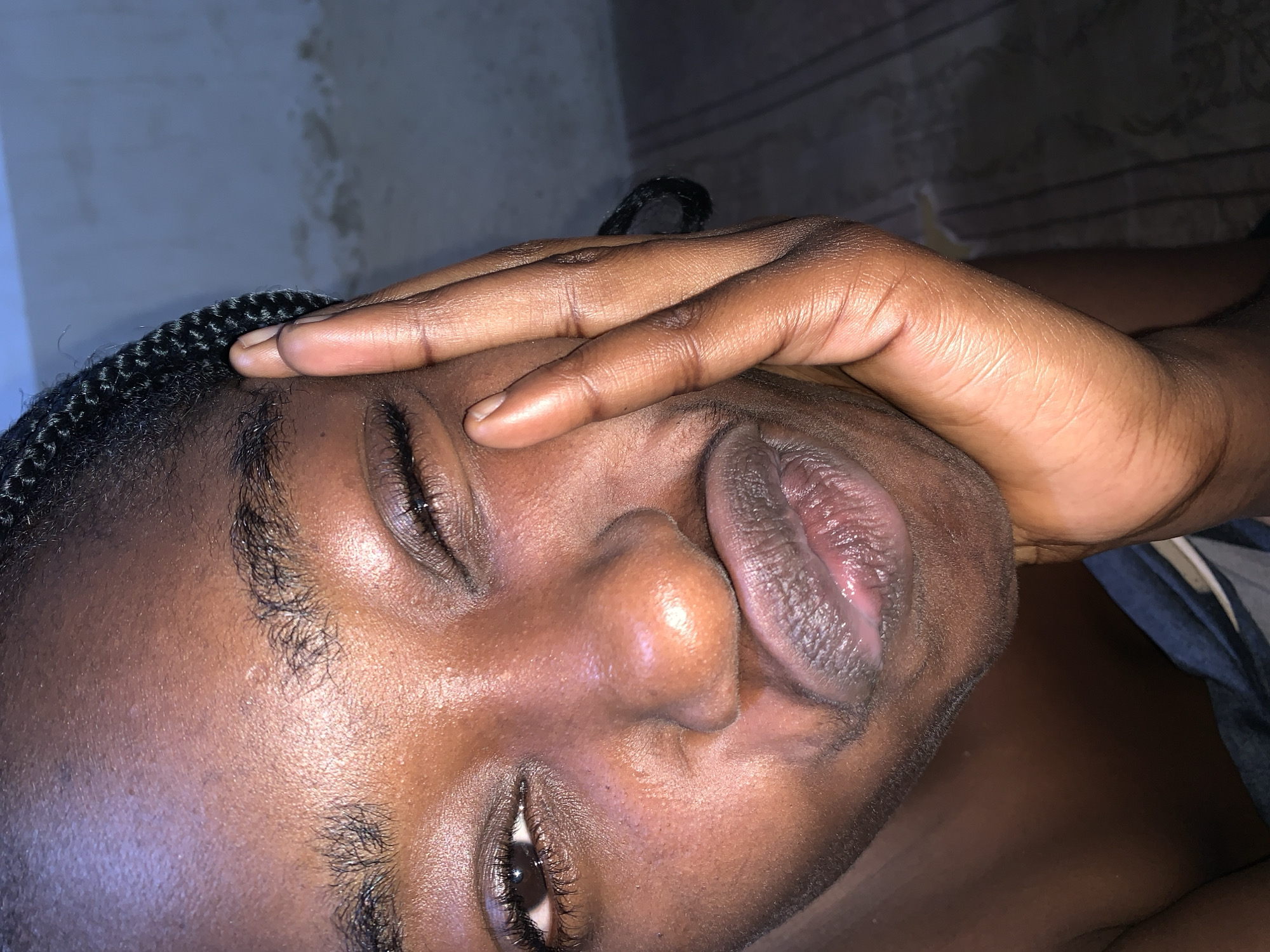 This is a close-up, head-and-shoulders portrait of a young Black person in Rafin Par, Nigeria. The subject's face is turned towards the camera, with their right hand resting on their cheek, obscuring part of their face and eye. Their lips are pursed, and their left eye is winking. Braided hair is visible on the left side of the frame, and a portion of dark fabric, possibly clothing, is visible at the bottom. The background is a dimly lit, textured wall and a patterned surface, suggesting an indoor setting. The lighting is warm and focused on the subject's face, creating highlights and shadows that emphasize the contours of their features. The image appears to have been taken in a residential setting within Rafin Par.