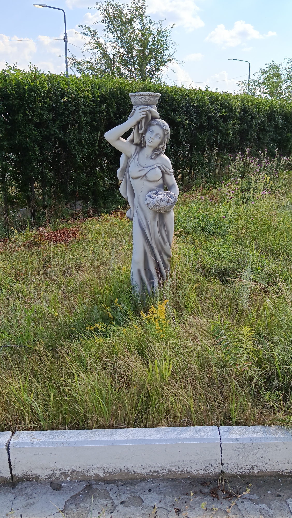 A full-shot, eye-level, outdoor medium portrait captures a gray, weathered stone statue of a woman standing in overgrown grass. The statue depicts a female figure with her hair pulled back, a draped garment covering her body, and a basket of flowers held in her left arm. She balances another basket on her head, with a cloth draped over her shoulder. The woman’s gaze is directed slightly upward and to her right, with a neutral expression.
Behind the statue, a dense, dark green hedge forms a wall, with a few trees visible above it. In the background, a bright blue sky with scattered white clouds is present. Two modern streetlights are visible on poles in the distance, one to the left and one to the right. The foreground is a patch of dry, yellowing grass and some wild yellow flowers. A cracked concrete curb runs along the bottom of the image, separating the grassy area from a paved surface. The overall impression is that of a neglected garden or public space. The location is identified as Kostanay, Kazakhstan.