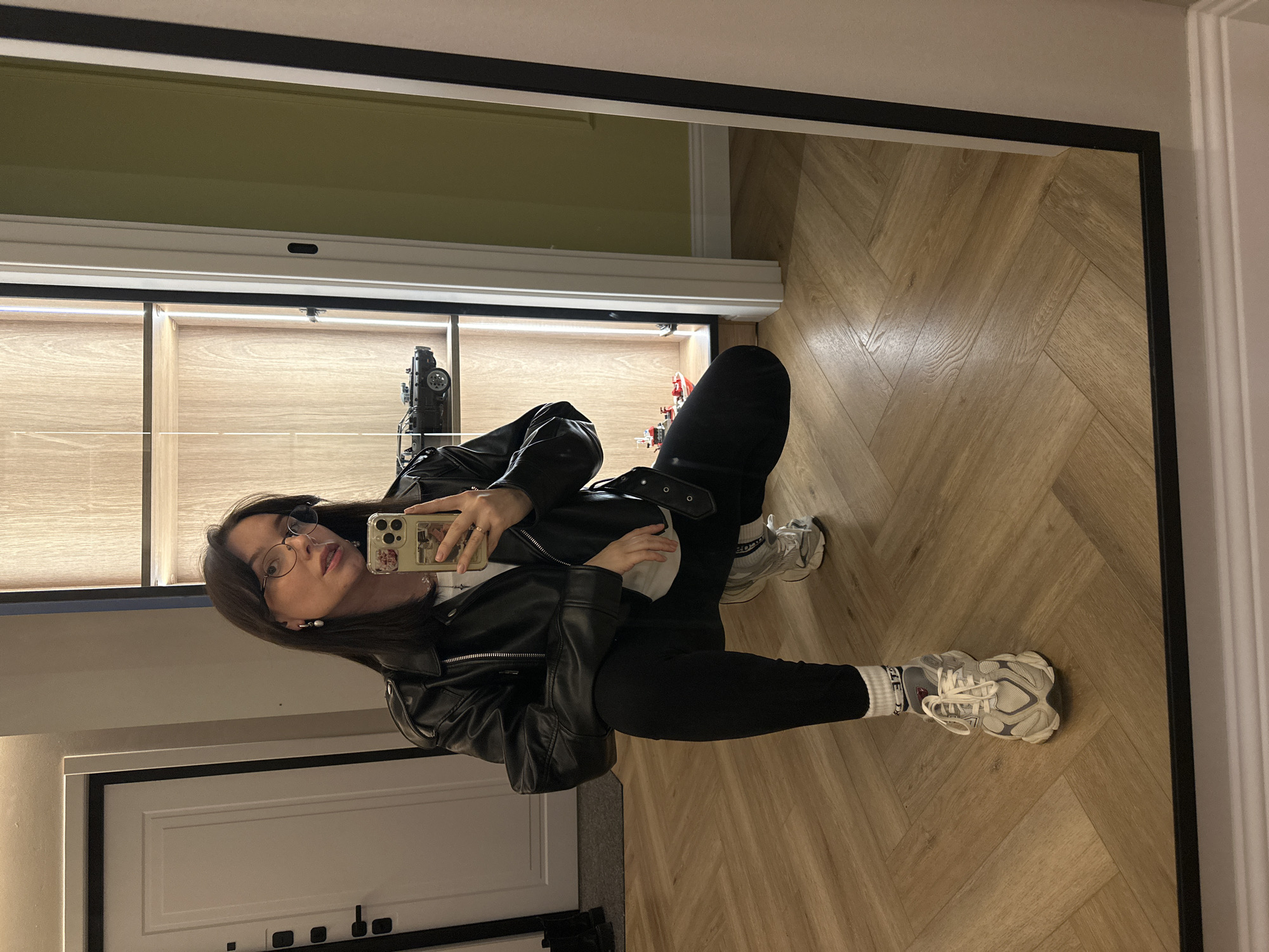 A person is seen in a mirror selfie. The person is wearing a black leather jacket, black pants, white socks, and sneakers. They are holding a cell phone. The reflection is of a hallway, with a wooden floor, a cabinet with a glass front, and a door. The location is potentially Chaikovsky, Russia, based on the user's request. No other details are available.
