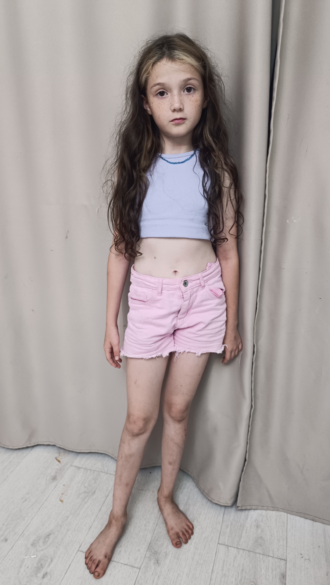 A young girl stands facing forward, leaning slightly against a backdrop of neutral-colored curtains. She has long, wavy brown hair with streaks of lighter brown, framing a face with light skin and freckles. She is wearing a light blue, ribbed crop top and pink denim shorts with frayed edges. Her bare feet are visible, appearing dirty, as are her lower legs. The floor beneath her is light-colored wood planks. The lighting is soft and even, suggesting an indoor setting, possibly a studio or room with diffused light. There is no visible text in the image. The girl's expression is neutral.