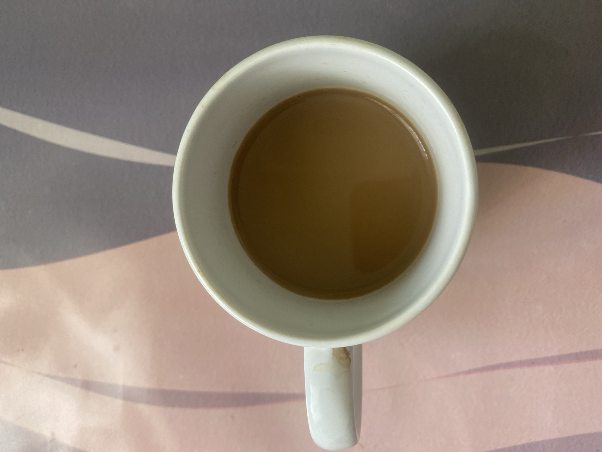 A top-down view of a white ceramic mug filled with a light brown liquid, likely coffee or tea with milk. The mug is sitting on a surface with a muted, patterned design in shades of grey and pink. A stain is visible on the mug's handle. The location in the city of Chaikovsky, Russia is not discernible from this image.