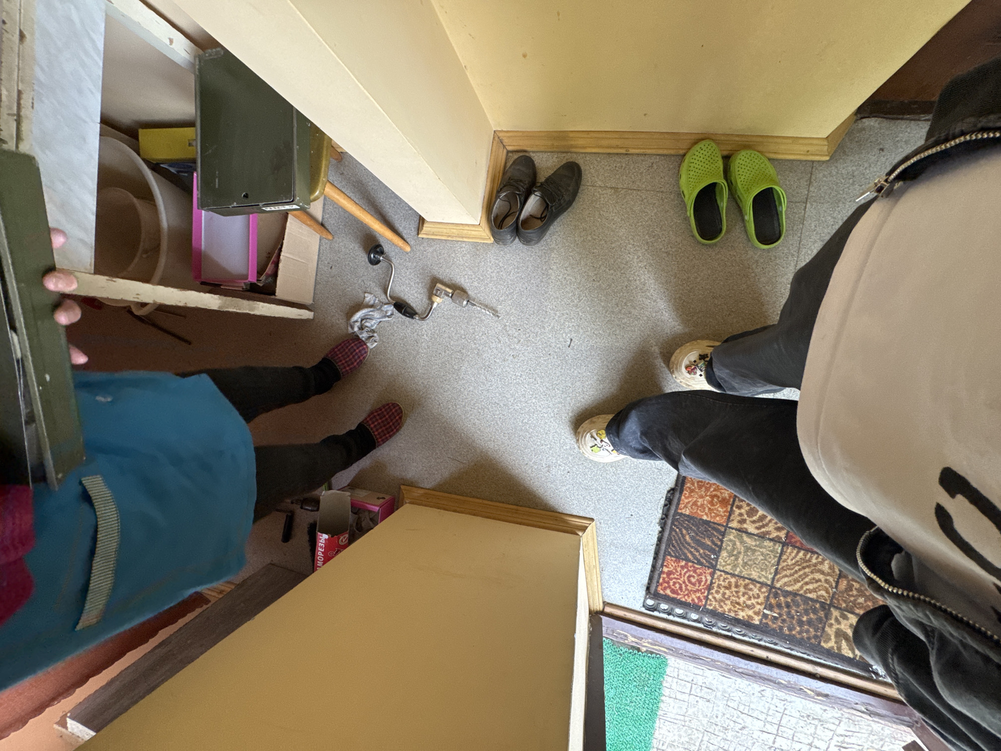 The image captures a group of people in an indoor setting, possibly a hallway or entryway, in Naro-Fominsk, Russia. The floor is a speckled gray, and the walls are painted a light, creamy yellow.
Several pairs of shoes are visible. On the left, a pair of dark shoes rests near a metal drill. To the right of those, a pair of vibrant green clogs is placed. Further to the right, a person is wearing light-colored sneakers with a colorful design on the soles, standing on a rug with a patchwork of brown, orange, and animal print squares. Another person is wearing plaid slippers with red and black checks.
In the background, a dark green metal box is on a shelf, next to a pink container. A wooden chair with a rounded back is also present. A wooden plank is lying on the floor in the foreground. Part of a blue garment, possibly a jacket or apron, is visible on the left side of the frame, along with a person's hand holding a green object. A small pink and red box with Russian text, "МОБЕЗЬЕ", is on the floor.
The overall scene suggests a casual, domestic environment. The lighting indicates it is likely daytime. There are no explicit clues about the weather or specific emotions of the individuals, but the presence of various footwear and tools might imply some activity related to home maintenance or a casual gathering.