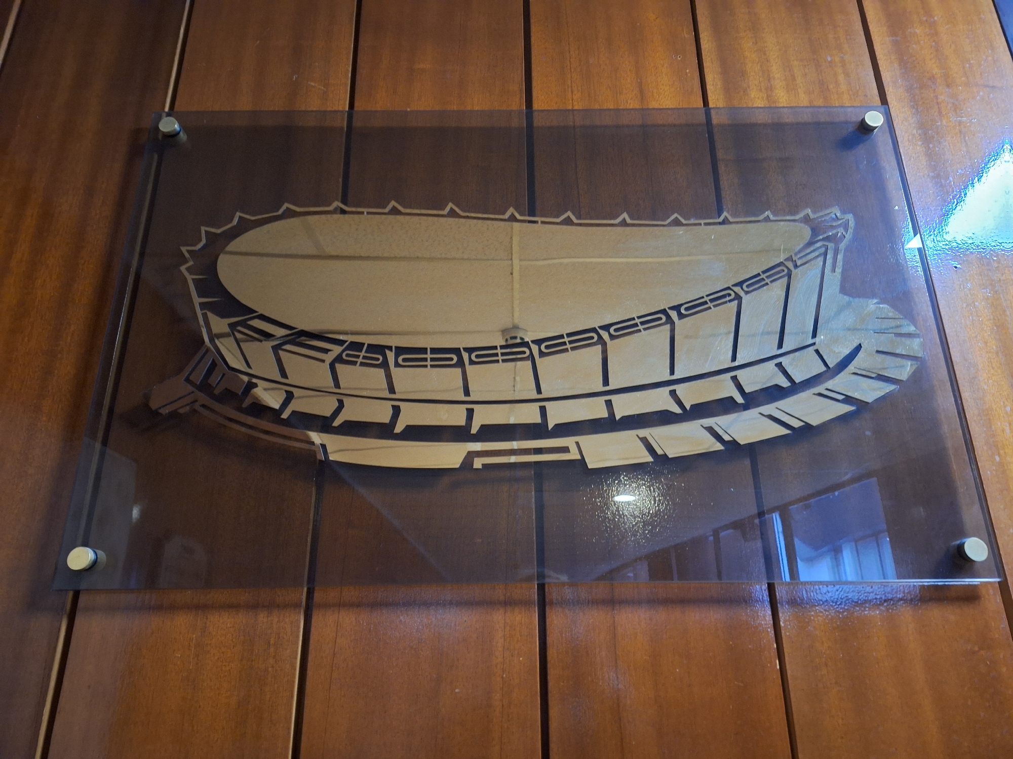 A photograph taken in Varna, Bulgaria, shows an architectural drawing of a stadium under a clear acrylic sheet, mounted on a wooden paneled wall. The drawing depicts an oval-shaped stadium with multiple tiers of seating. The top edge of the stadium is lined with a series of triangular points. The drawing is a light color and appears to be laser cut. The acrylic sheet is fixed to the wall with four silver-colored standoffs, one at each corner. Reflections on the surface of the acrylic suggest a window or light source nearby. The wooden panels on the wall are vertically aligned and stained a medium brown color.