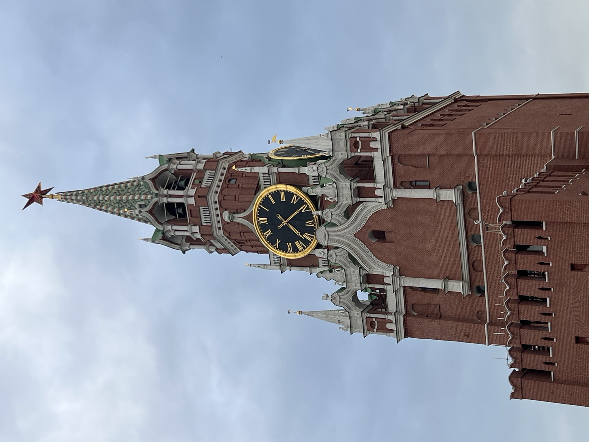 The image shows a low-angle view of the Spasskaya Tower of the Moscow Kremlin. The tower is made of red brick and features a green spire with a red star on top. A large clock face is visible on the tower, with Roman numerals and gold trim. The sky is overcast and gray.