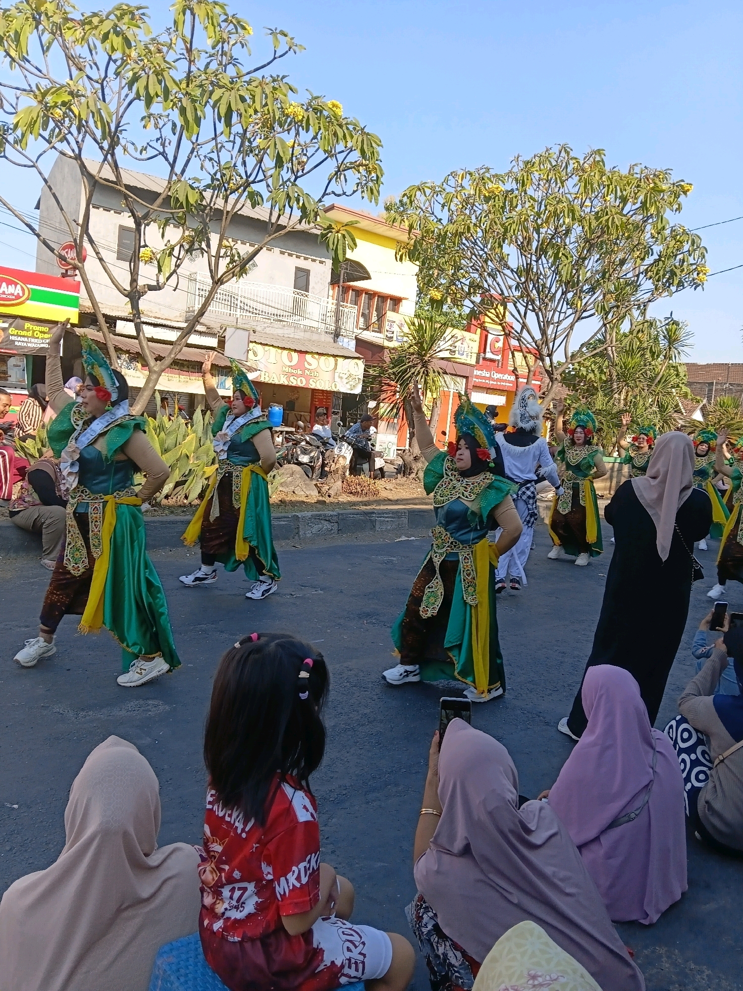 A group of dancers in vibrant, traditional costumes are performing in the middle of a street in Wadungasri, Indonesia. The dancers, adorned with green and gold garments, elaborate headpieces, and red floral accents, move in unison with raised arms. Behind them, buildings line the street, some painted in bright yellow and others in muted tones. Trees with green foliage and yellow flowers are scattered throughout the scene, suggesting a warm, sunny day. On the sidewalks and road, spectators, including women in hijabs and children, are seated or standing, some capturing the performance with their mobile phones. The atmosphere appears festive, with a cultural performance taking place in a semi-urban setting.