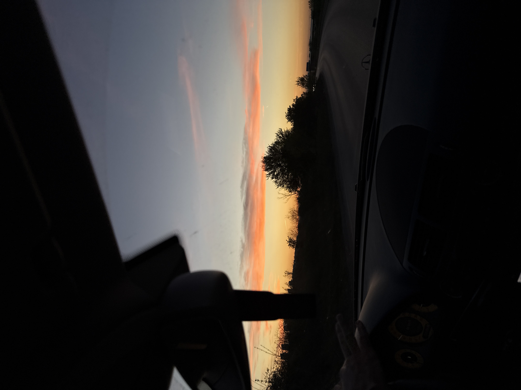 The media file is a wide-angle shot taken from inside a car, looking out at a scenic sunset or sunrise. The sky dominates the upper portion of the image, showcasing a gradient from pale blue/grey at the top to vibrant orange and yellow closer to the horizon. Wispy, elongated clouds, tinged with pink and orange, stretch horizontally across the sky.
Along the horizon, a dark silhouette of trees and sparse vegetation is visible, suggesting a rural or open landscape. A road curves along the right side of the frame, disappearing into the distance.
The car's interior frames the scene, with dark areas of the dashboard, windshield, and side pillars visible on both the left and right sides. In the lower right corner, a hand is discernible, resting on or near the dashboard, where circular, unreadable gauges or controls are faintly visible in the deep shadows.
The time of day is clearly dawn or dusk, indicated by the colorful sky. The weather appears clear with light cloud cover. The overall mood is serene and captures a fleeting moment of natural beauty from a moving or stationary vehicle. The location context indicates this scene is near Sofia, Bulgaria. No visible text is present in the image.