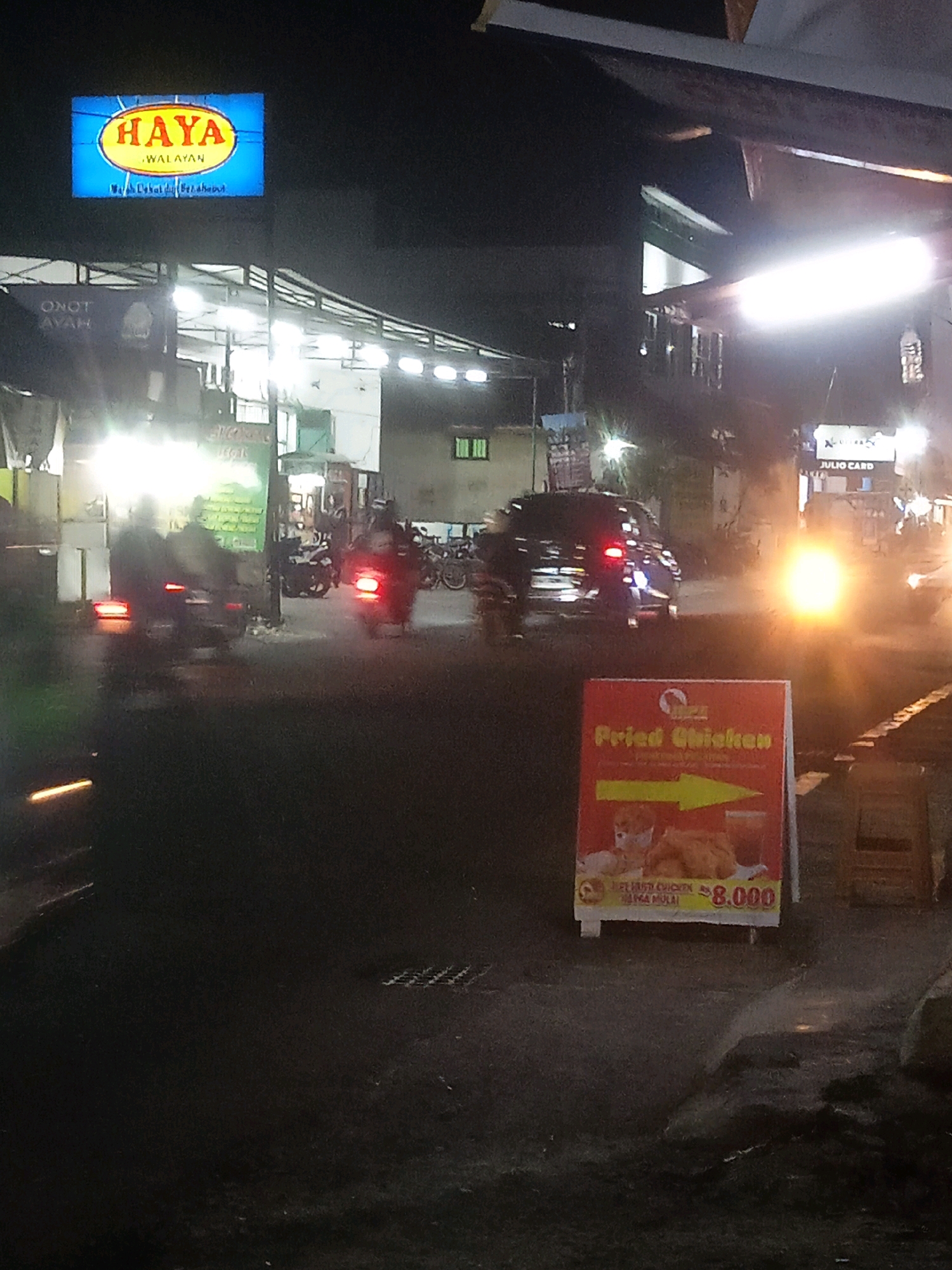 The image shows a nighttime street scene in Trihanggo, Indonesia. A brightly lit blue and yellow sign for "HAYA WALAYAN" is visible, along with other illuminated signs. Several motorcycles are on the road, with some appearing to be in motion and others parked. A dark-colored car is also present. In the foreground, a red sign advertises "Fried Chicken" with a yellow arrow pointing to the right and the price "Rp. 8.000". The environment appears to be a commercial or roadside area with buildings and streetlights contributing to the illumination. The overall scene suggests a lively, active urban environment at night.