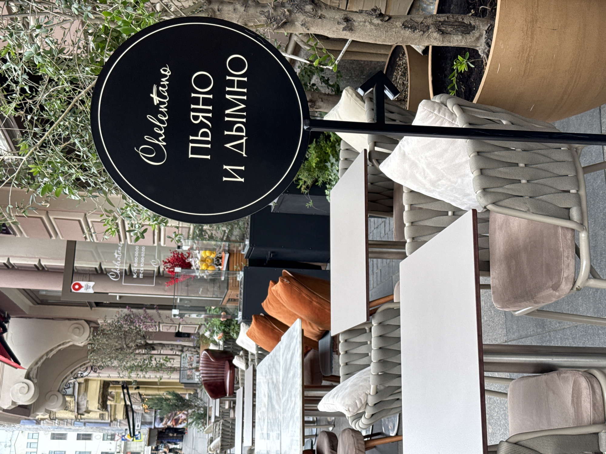 The image shows an outdoor seating area of a restaurant called "Chelentano," as evidenced by the circular black sign with white lettering bearing the restaurant's name in both Latin and Cyrillic scripts ("Chelentano" and "ПЬЯНО И ДЫМНО" respectively). The location is identified as Saint Petersburg, Russia by the presence of Cyrillic text and the architectural style of the surrounding buildings.
The scene is primarily composed of tables and chairs set up for outdoor dining. Each table has a white top and is accompanied by chairs with woven backs and seats in neutral tones, some with cushions. The chairs are arranged neatly, suggesting the area is ready for customers.
In the background, the streetscape of Saint Petersburg is visible, showing buildings with decorative facades and awnings. A sign visible in the background lists the restaurant's hours of operation in Cyrillic. Patrons and activity are not evident in the immediate foreground, though blurred figures can be seen in the distance. Additionally, greenery in planters adds a touch of nature to the urban setting.