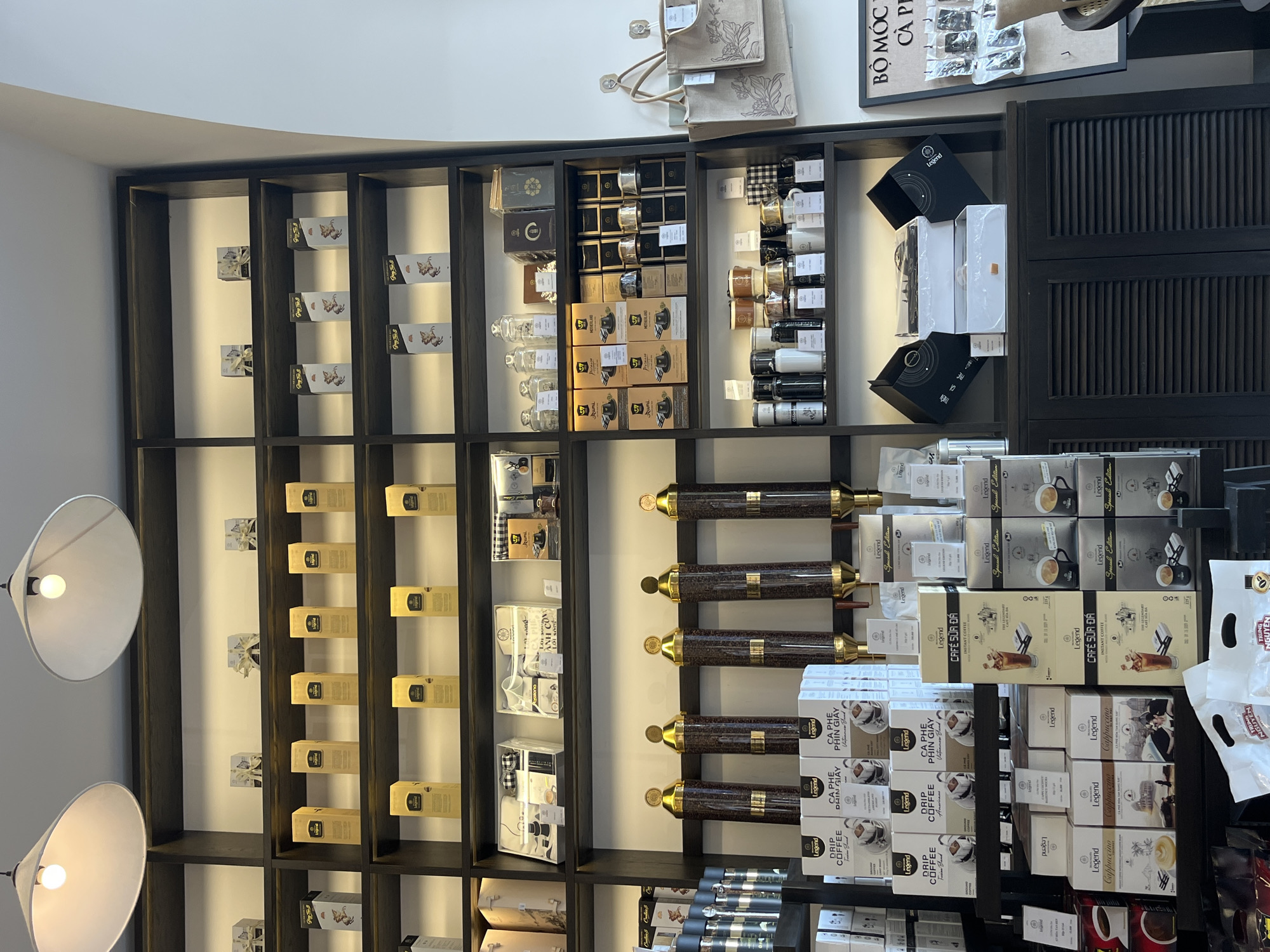 The image shows a retail space in Nha Trang Ward, Vietnam, featuring a dark wooden shelving unit displaying coffee products. The shelves are arranged vertically and horizontally, showcasing various coffee brands and types, including "Legend" and "Rumi." Products include drip coffee, instant coffee, and coffee accessories such as filters and brewing devices. Some shelves contain decorative items and bags. The lighting in the space is provided by lamps with large, round shades. A sign is visible with text that may include the words “BỘ MÓC CÀ PH.” The background is a light-colored wall.
