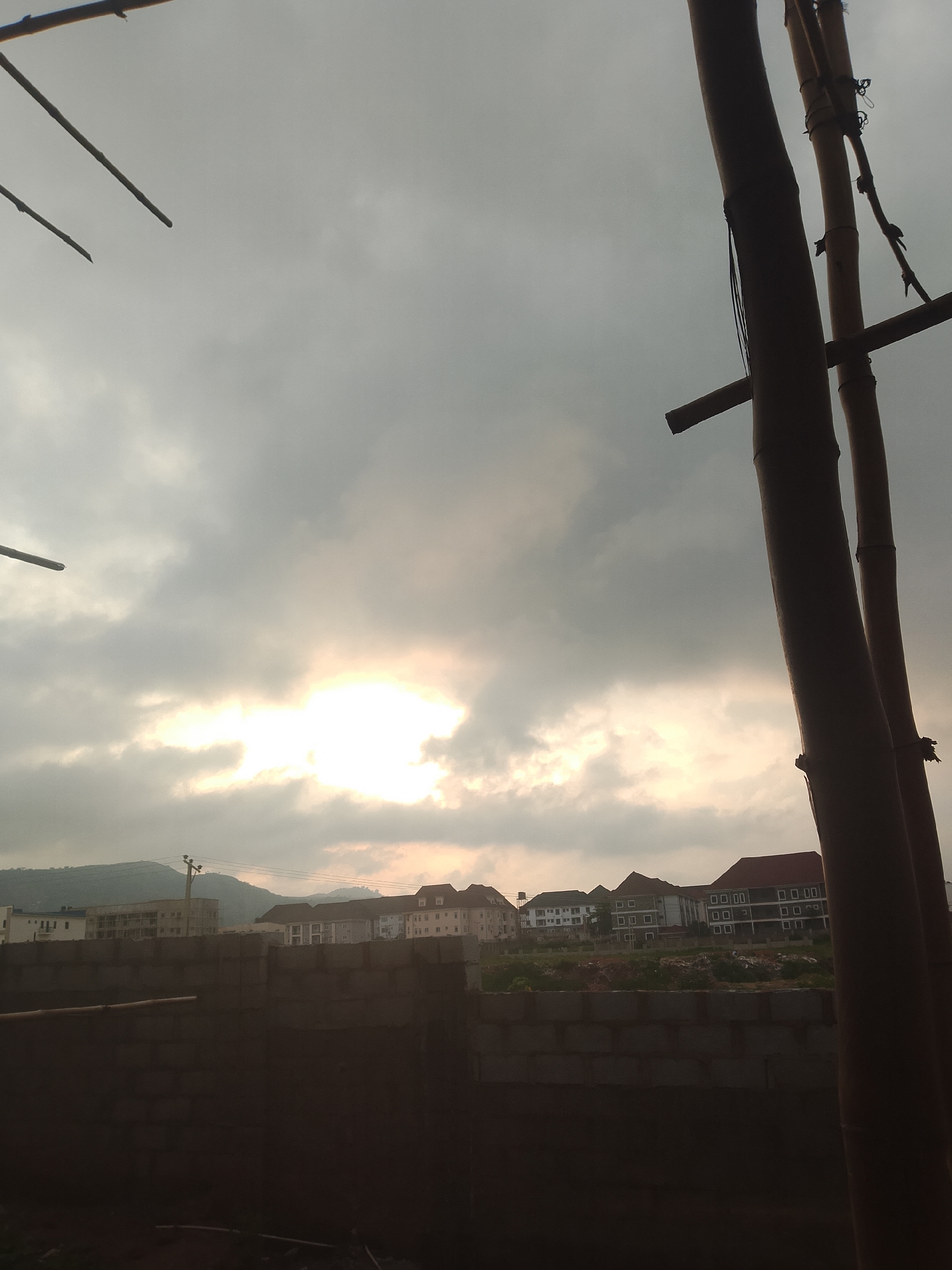 The image shows a cloudy sky with the sun breaking through, casting a bright light in the center. In the foreground, there is a partially constructed brick wall. Beyond the wall, there are several modern buildings with multiple stories and windows. In the background, a hilly landscape is visible. The scene appears to be a construction site in a semi-urban or suburban area, possibly on the outskirts of a city. The overall lighting suggests it is either early morning or late afternoon.