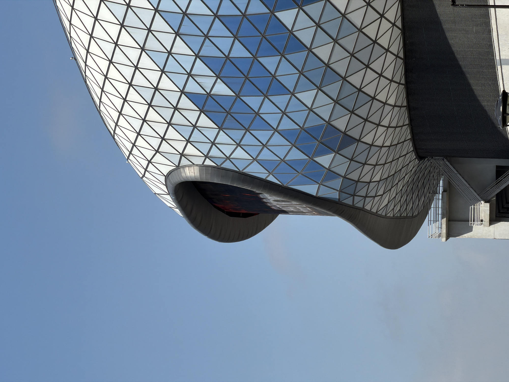 The image shows a portion of the Moscow Planetarium building in Moscow, Russia. The structure features a dome with a geodesic pattern of triangular glass panels that reflect the sky. The lower portion of the structure has a curved, modern design. A blue sky fills the background. The photograph is oriented with the building appearing to reach upwards.