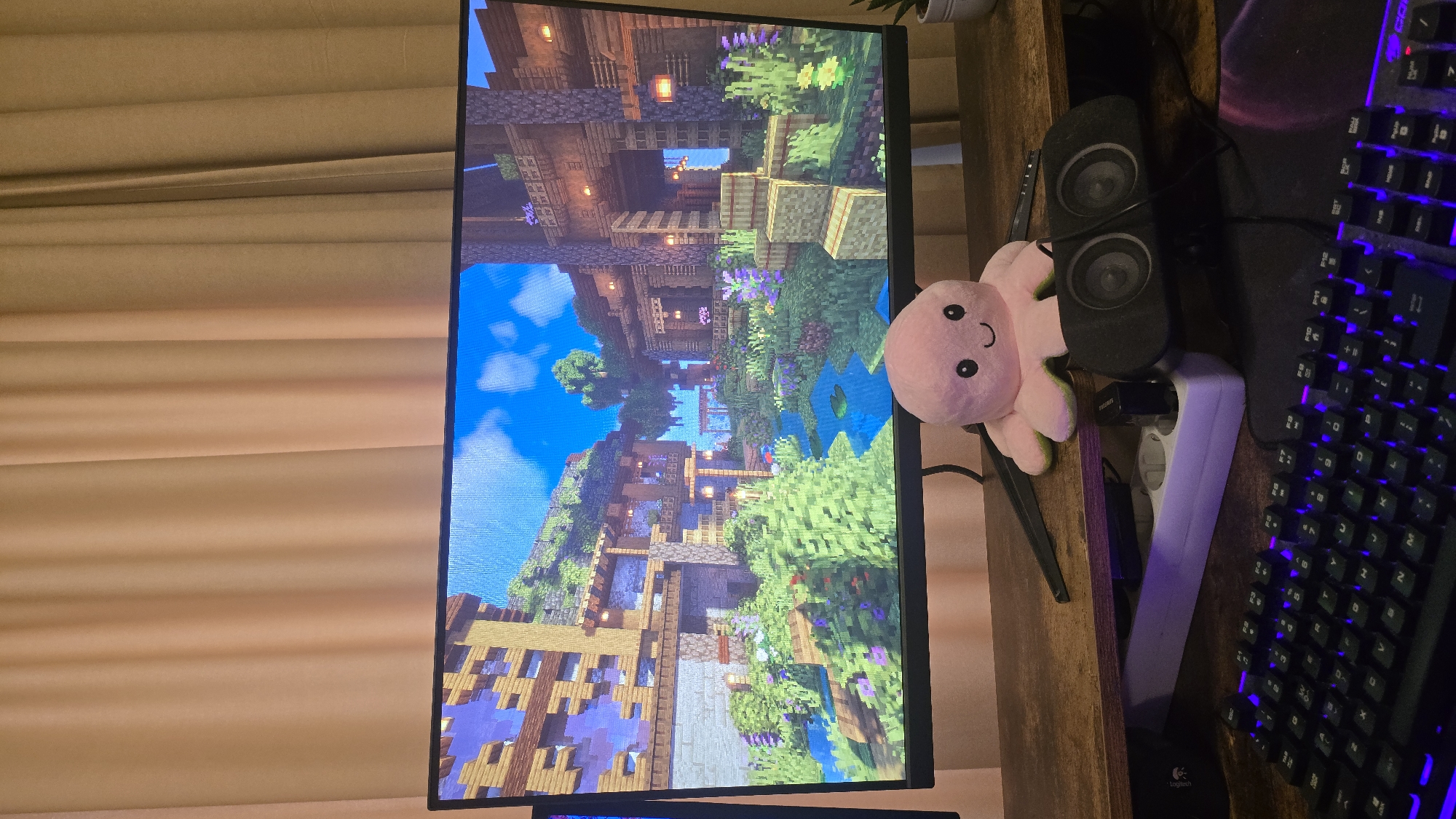 This vertical image captures a personal computer setup on a wooden desk, likely in Sofia, Bulgaria, characterized by gaming peripherals and decorative elements, set against light-colored window blinds.
The central focus is a computer monitor displaying a vibrant, detailed video game scene, strongly resembling Minecraft. The game world features intricate multi-level wooden structures with glowing lanterns, lush green foliage, various purple and yellow flowers, a pond with lily pads, and a bright blue sky with scattered clouds. The architectural style suggests an elaborate, custom-built environment within the game.
To the right of the monitor, on the wooden desk, sits a pink plush octopus toy with a friendly, smiley face. Adjacent to the octopus are two black computer speakers stacked vertically. Further to the right, a black mechanical keyboard with distinctive purple-blue backlighting is visible, resting on a dark mouse pad which faintly displays "Logitech" near the bottom edge. A white power strip is positioned between the speakers and the keyboard, with various cables visible behind the peripherals. A small portion of a green potted plant can be seen at the very top right corner of the frame.
The left side of the image is dominated by light-beige horizontal window blinds or curtains, suggesting an indoor setting and providing ambient light. The overall lighting implies it could be daytime, though the room itself appears dimly lit, possibly due to the blinds being closed or it being evening, with the monitor providing the brightest light source. No human activity is depicted, but the setup indicates a space for gaming or computer use.