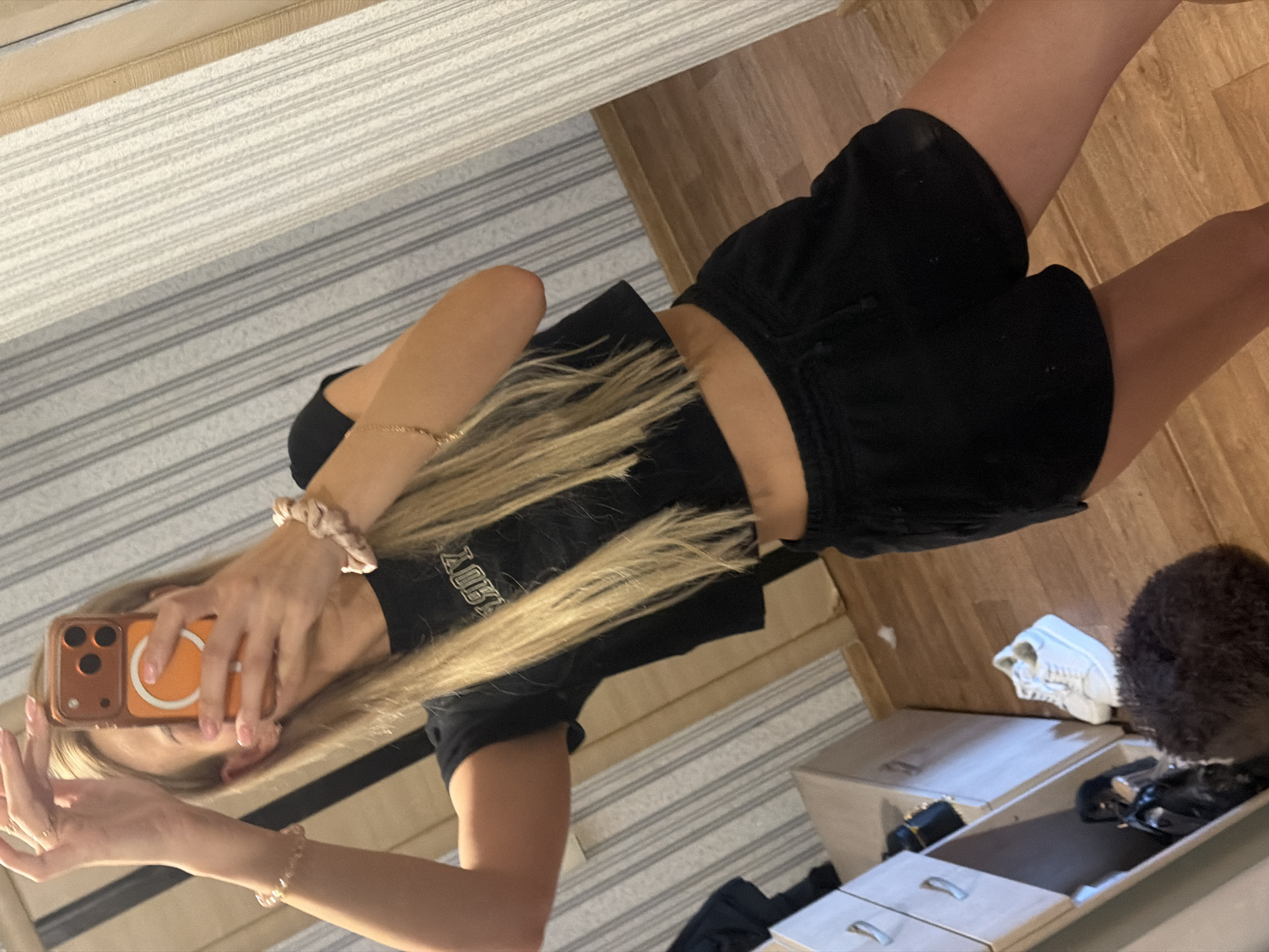 A young woman with blonde hair is taking a mirror selfie in what appears to be a bedroom. She is wearing a black crop top and black shorts. On her left wrist, she has a bracelet made of pink beads, and on her right wrist, she has a scrunchie. She is holding her phone in front of her face with both hands, obscuring her face. The background shows a striped wallpaper, a dresser with drawers, and a wooden floor. There are white sneakers on the floor next to the dresser, and a dark, curly object that may be a rug or a pet. The scene is indoors and appears to be a domestic setting. There is no information to suggest this location is in Kyiv, Ukraine.