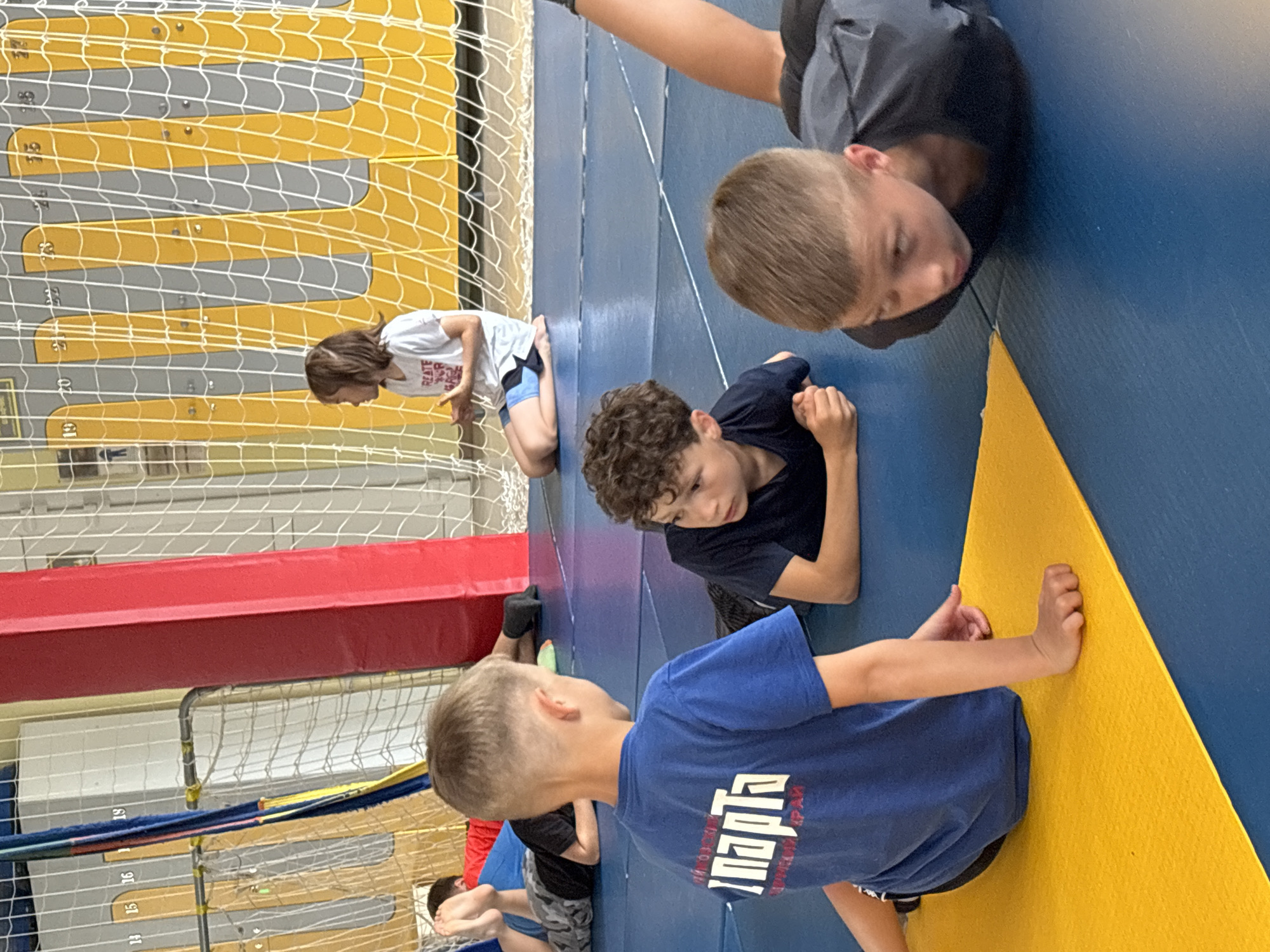 This image depicts an indoor sports training session involving several children, presumably boys, in a gym located in Chaikovsky, Russia. The setting features a floor covered with large blue mats, and a background wall paneled with alternating yellow and grey padding, marked with numbers ranging from 19 to 26. A white net is stretched across parts of the background, and a red padded beam is also visible, possibly covering a support structure.
In the foreground and midground, four children are prominently featured, all lying on their stomachs on the blue mats. The child closest to the camera, with short light brown hair, wears a blue t-shirt featuring white Cyrillic text, which clearly reads "СПАРТА" (SPARTA), indicative of a sports club or team. He is looking towards a yellow mat at the edge of the floor. Adjacent to him, a boy with curly hair in a dark t-shirt is also lying prone, looking down. Further back, another child in a dark grey t-shirt lies similarly. To the left, a fourth child in a white t-shirt and blue shorts is kneeling against the padded wall.
The children's postures suggest they are engaged in a physical conditioning exercise, possibly a warm-up, cool-down, or a specific drill related to wrestling or grappling. Their expressions are generally focused and neutral, indicating concentration. The indoor environment is brightly lit, likely by artificial lighting. The Cyrillic text on the visible t-shirt, combined with the provided location context, confirms the setting in Russia.