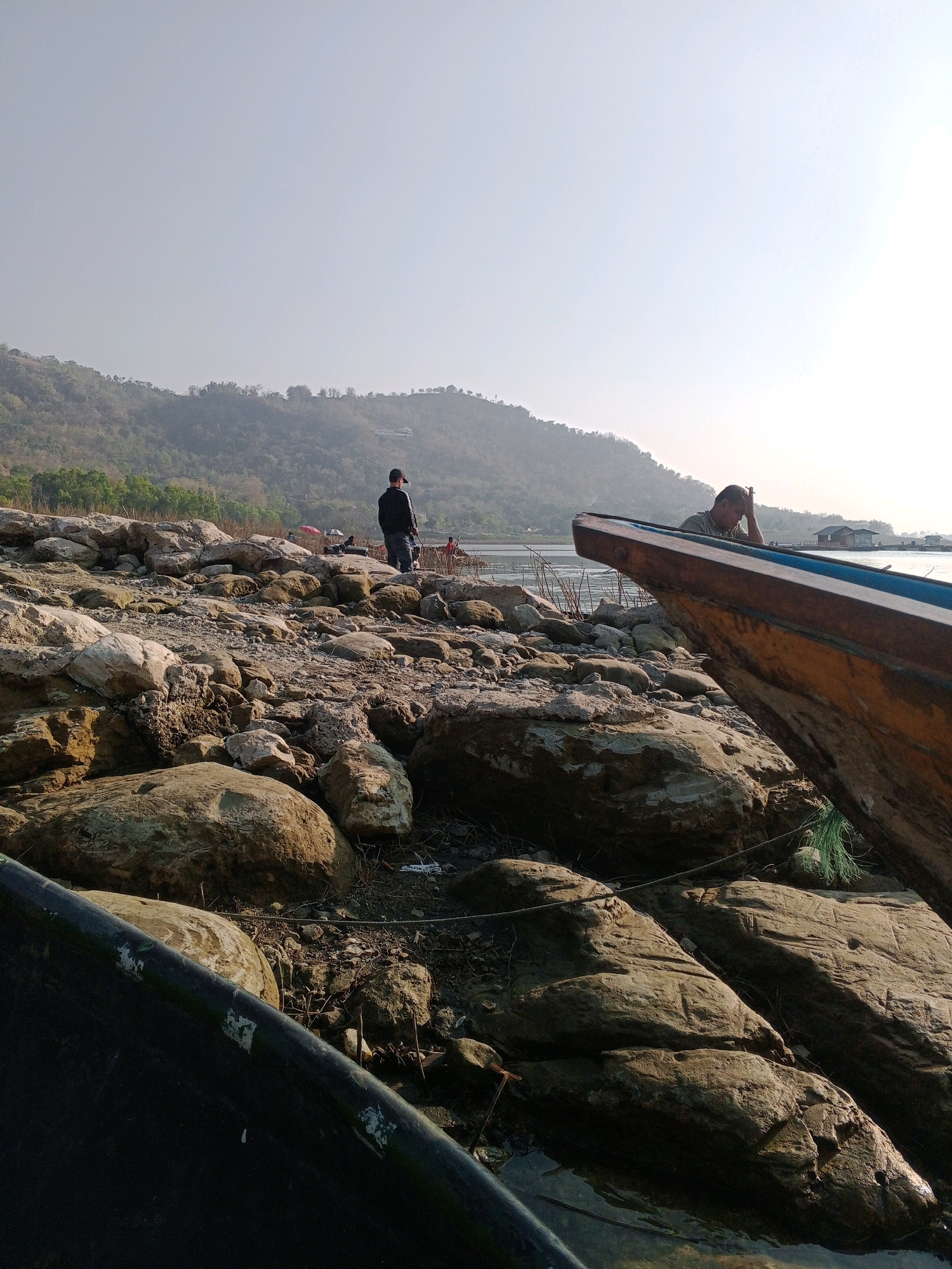 This image depicts a rocky shoreline with a body of water in the background. In the mid-ground, a person wearing a dark shirt and a baseball cap stands facing away from the camera, looking towards the water. Further in the background, a hilly landscape is visible under a hazy sky. In the foreground, the edge of a wooden boat is prominent, with another boat visible in the far distance with a person inside. The scene appears to be in Sendang, Indonesia, given the presence of a boat and typical landscape elements.
