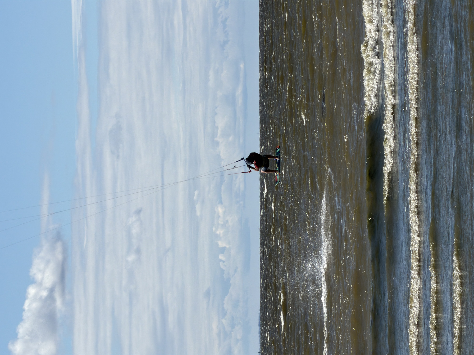 A person is kitesurfing on a body of water under a blue sky with scattered clouds. The kitesurfer is wearing a dark shirt and shorts, and is crouched on a board. The water is choppy with waves, and there is a spray of water near the surfer. The horizon line is visible in the distance, with land or trees appearing as a faint line. The image does not provide any specific indicators of the location being Saint Petersburg, Russia.