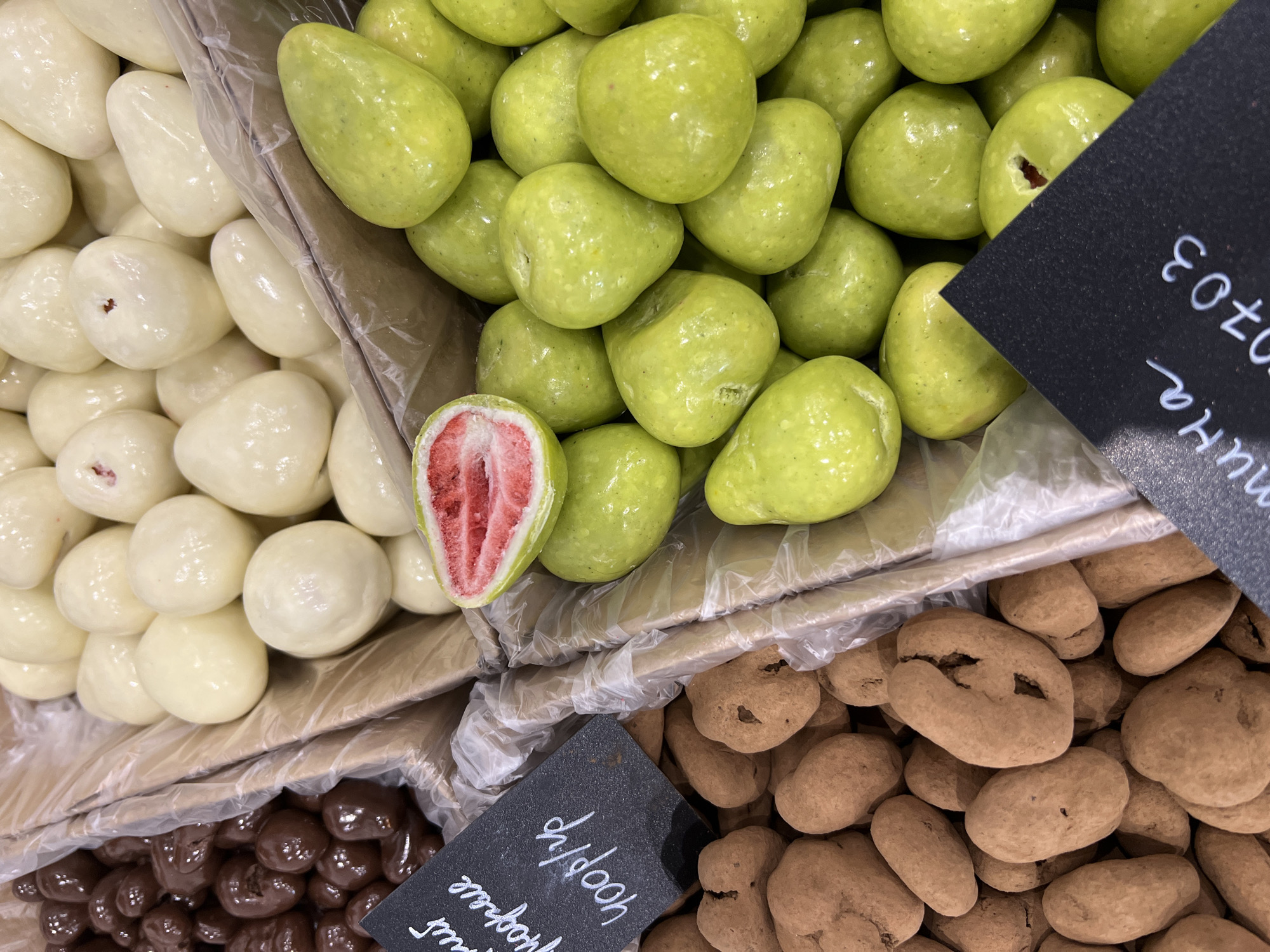 The image shows a display of chocolate-covered fruits in a store in an unknown city. The fruits are arranged in four sections within a cardboard container lined with plastic. The top left section features white chocolate-covered fruits, the top right contains light green chocolate-covered fruits, the bottom left section contains dark brown chocolate-covered fruits, and the bottom right contains brown chocolate-covered fruits coated in a dusty brown powder. A cut strawberry with a red interior is visible among the white and green fruits. Two small black cards with white handwriting are positioned near the bottom right and top right sections. The handwriting on the cards is not readily decipherable.