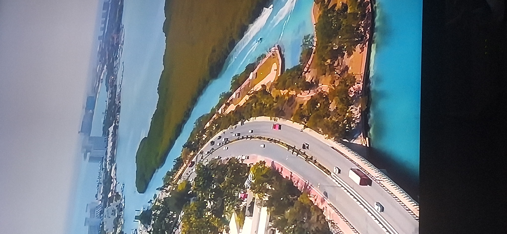 An aerial view shows a coastal city with a highway bridge spanning over turquoise water. The bridge is populated with vehicles, including a red truck and several cars. Along the left side of the highway, there are trees and buildings. To the right of the highway, a body of water is visible, with a small boat and a larger boat leaving a wake. In the distance, more buildings and water can be seen, with a green forested area separating the highway from the larger body of water. The sky is hazy and overcast. This image does not depict Bila Tserkva, Ukraine, as the geography and architecture suggest a tropical or subtropical coastal location.