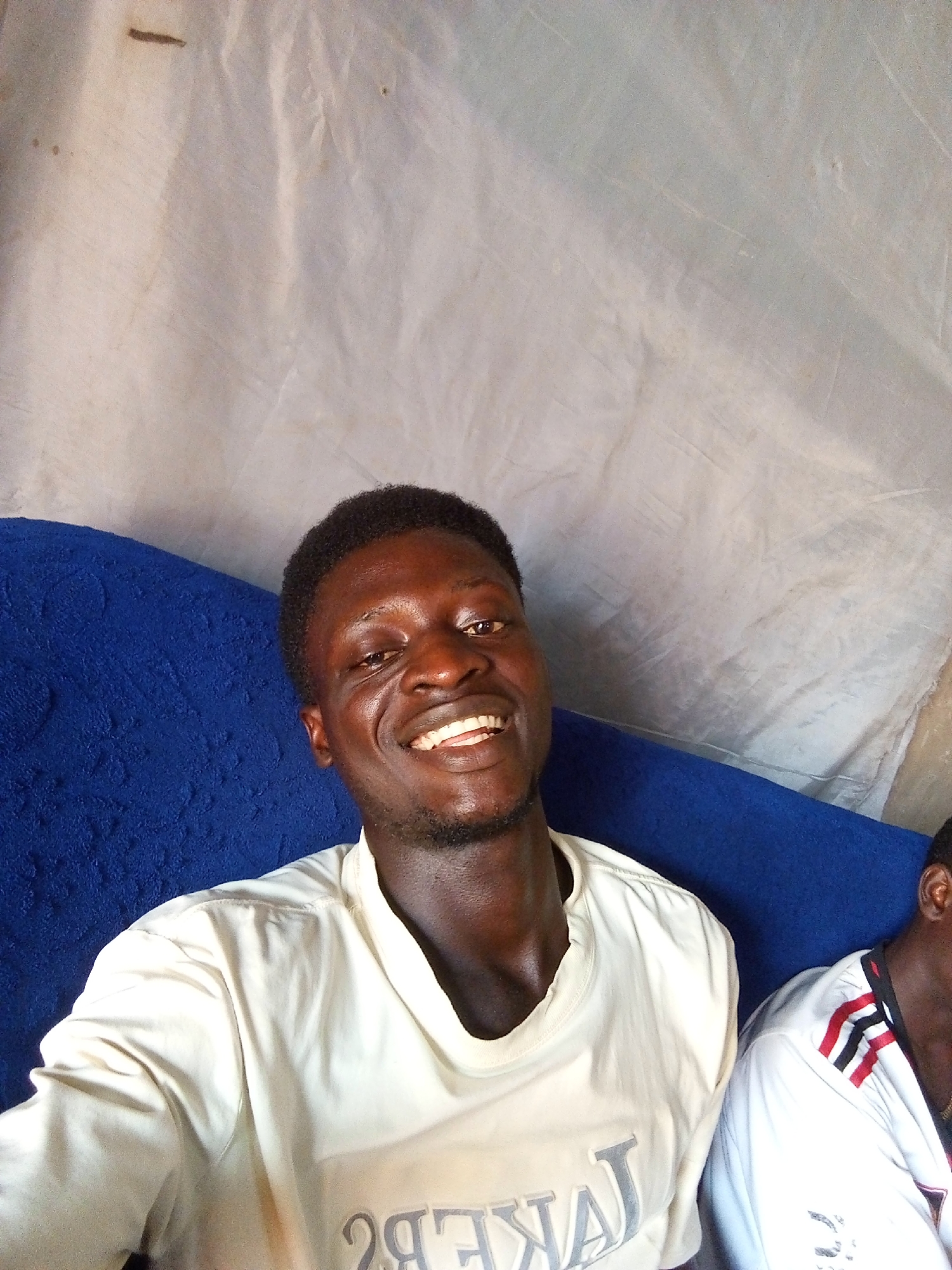 A close-up selfie shows a Black man with dark skin, short black hair, and a beard, smiling directly at the camera. He is wearing a cream-colored long-sleeved shirt with "LAKERS" printed in a light gray, stylized font across the chest. He is seated on a blue textured surface, possibly a couch or cushion. In the background, a light-colored, crumpled fabric is visible, along with another person wearing a white shirt with red and black stripes on the collar, partially visible to the right. The setting appears to be indoors, with a simple, possibly temporary, backdrop. There are no discernible landmarks or contextual clues to suggest a specific location within Nyabe, Nigeria.