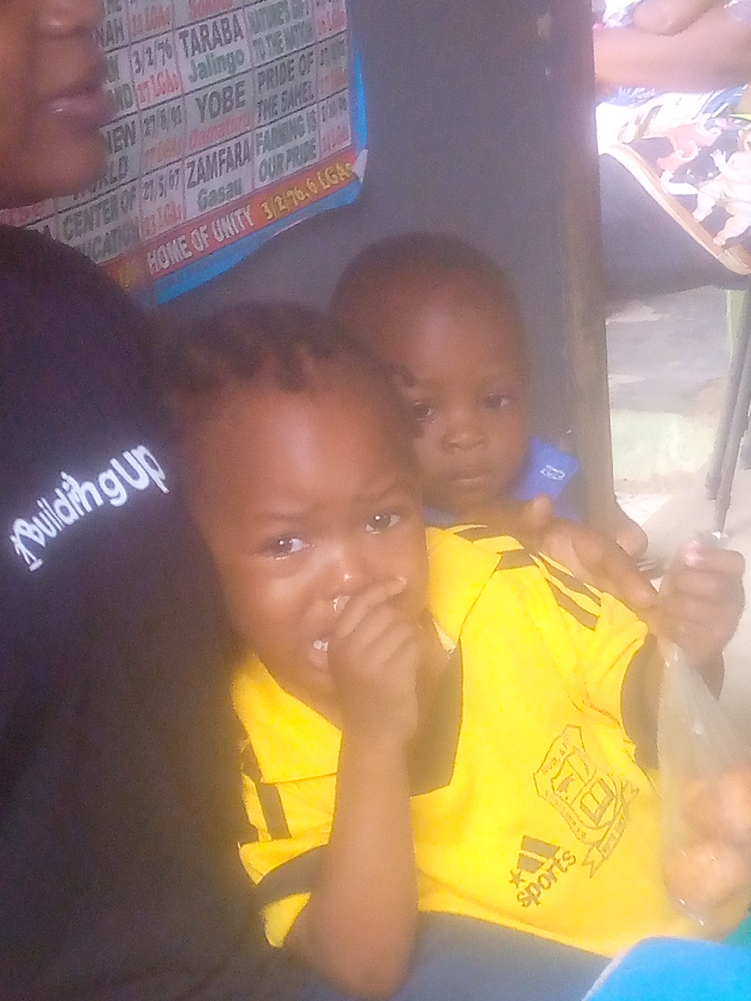 Two young children are in the frame. The child in the front is wearing a yellow Adidas shirt and appears to have their fingers in their mouth. Behind the first child, partially obscured, is another child wearing a blue shirt with an unidentifiable logo. The context of the location in Ikorodu, Nigeria, is supported by text visible on a sign behind the children, referencing Nigerian states (Taraba, Yobe, Zamfara) and the phrase "HOME OF UNITY 3/2/76.6 LGA," as well as the word "EDUCATION." A person wearing a black shirt with the text "Building UP" is on the left. There is a table next to the children, and it is not clear if they are sitting on or near it.