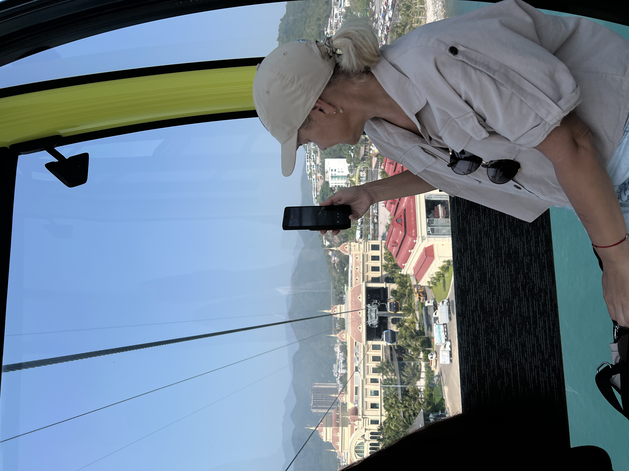 A woman is inside a cable car, taking a photo with her smartphone of the view below. She's wearing a light-colored shirt, a baseball cap, and sunglasses are tucked into her shirt. The cable car is traveling over the city of Nha Trang, Vietnam, which is visible through the window. Mountains are in the background. The cable car's cable is also visible through the window. A large building complex, with a red-tiled roof, is directly below the cable car. Palm trees dot the landscape below. The interior of the cable car is a mix of yellow and black.