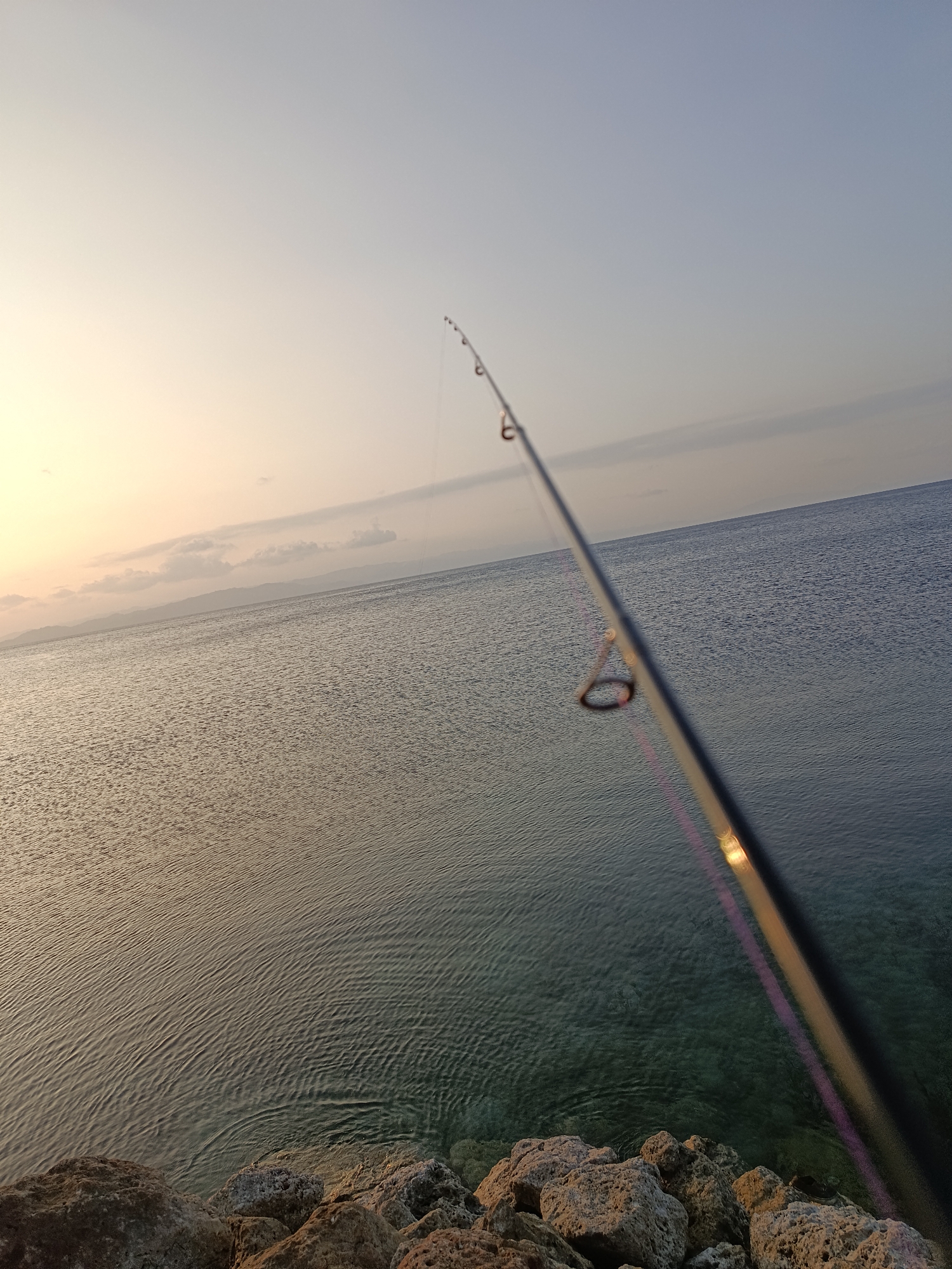 The image shows a fishing rod extending into the water. The rod is black with several guides, and a fishing line is visible. The water is calm with gentle ripples, and the sun is setting in the background, casting a warm glow on the scene. The shoreline is rocky, with several large stones in the foreground. The sky is a soft gradient of pale yellow and blue, with a few wispy clouds. The image appears to have been taken at sunrise or sunset. The location is identified as Paisulamo, Indonesia, which is a coastal village. The overall mood of the image is peaceful and serene.