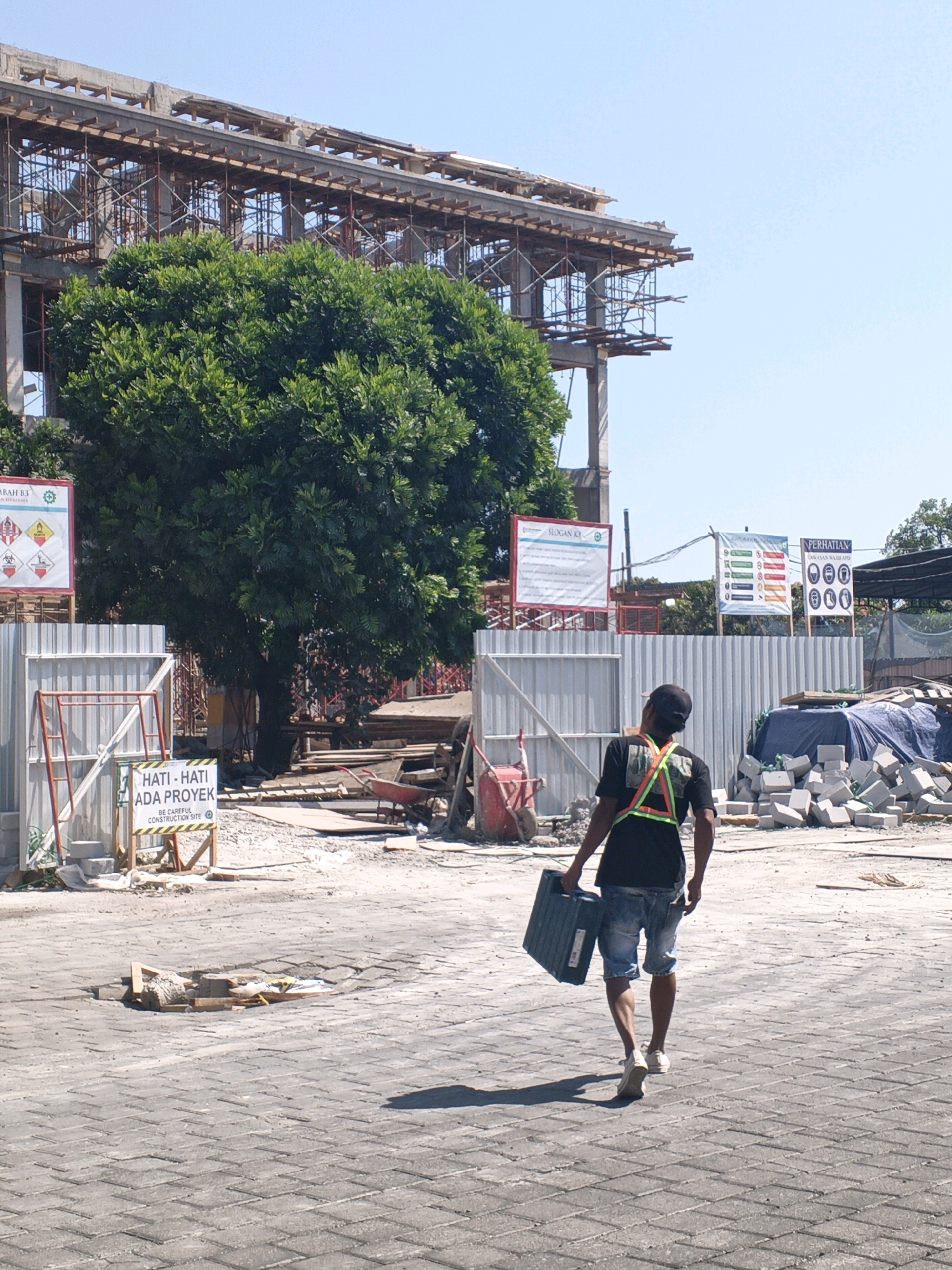 The image depicts a construction site in Denpasar, Indonesia, under a clear blue sky. In the foreground, a construction worker, seen from behind, walks away from the camera carrying a dark green toolbox. He wears a black t-shirt, denim shorts, and a high-visibility green and orange vest. His legs are visible from the knees down, showing light-colored sneakers. The ground is paved with gray interlocking bricks, with some scattered debris, including wooden planks and construction materials, in the mid-ground.
Behind the worker, a large, lush green tree dominates the mid-ground, partially obscuring a partially constructed building. The building features concrete structures and extensive scaffolding made of metal poles and wooden planks. Several signs are visible on metal fences surrounding the site. One sign in the foreground, with a yellow border and red accents, reads "HATI-HATI ADA PROYEK" and "BE CAREFUL CONSTRUCTION SITE". Further back, other signs with various symbols and text, some related to safety regulations and slogans, are attached to fences.
In the background, more construction materials, including stacks of grey bricks and blue tarps covering what appear to be building materials, are visible. A red wheelbarrow is positioned near a pile of wooden planks. The overall scene suggests an active construction project in an urban or semi-urban environment.