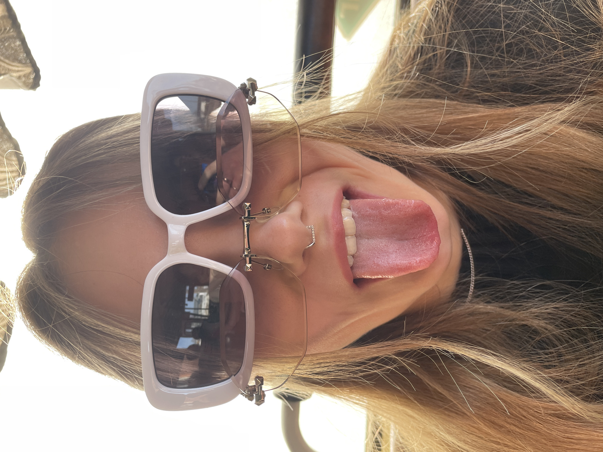 A close-up portrait depicts a female subject from the chest upwards, centered in the frame. The subject has long, light brown hair, appearing somewhat disheveled. She is wearing two pairs of glasses: a primary pair of large, white-framed rectangular sunglasses covering her eyes, and a secondary pair of smaller, rimless glasses with thin gold-toned frames positioned beneath the sunglasses. A small, circular, silver-colored nose ring is visible on her left nostril. Her mouth is open, with her tongue fully extended outwards and downwards. A thin, silver-colored chain necklace is visible around her neck, above a dark garment. The background is overexposed with bright light, suggesting strong backlighting or an outdoor setting. Reflections in the sunglasses show indistinct shapes, possibly indicating an interior environment like a vehicle cabin. The specific location within the city of Фоки, Russia, cannot be determined from the visual content of the image.