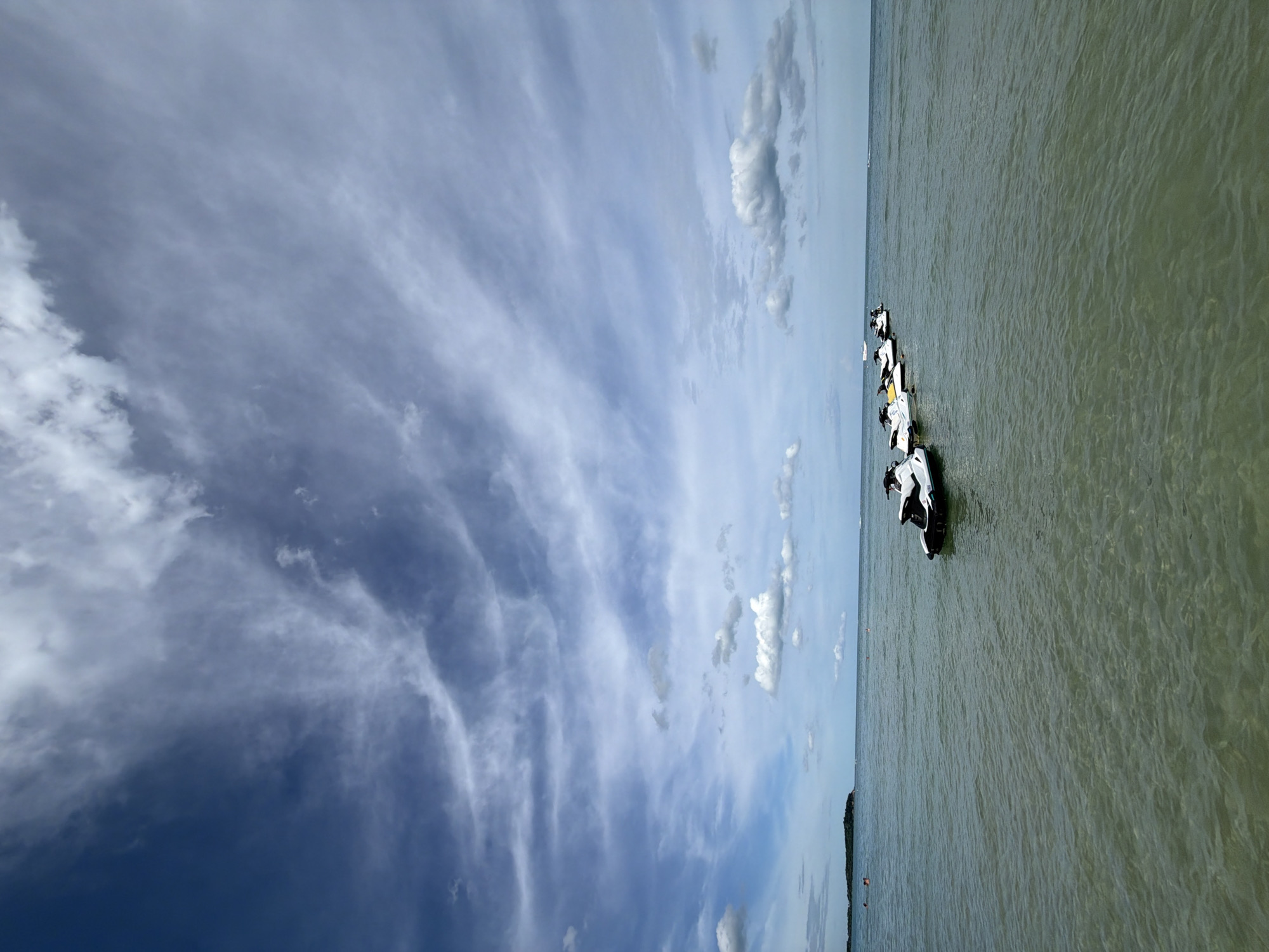 The image shows a line of jet skis moored in the shallow waters of Baan Chaweng, Thailand. The sky is filled with dramatic, wispy clouds against a blue backdrop, hinting at a dynamic weather system. The water is calm and clear, with sunlight creating patterns on the sandy bottom. In the distance, a small, tree-covered island is visible on the left, and a few people can be seen swimming or wading in the sea. The jet skis, which appear to be mostly white with black accents, are arranged in a diagonal line extending from the lower left to the middle right of the frame. The overall scene evokes a sense of leisure and outdoor recreation.