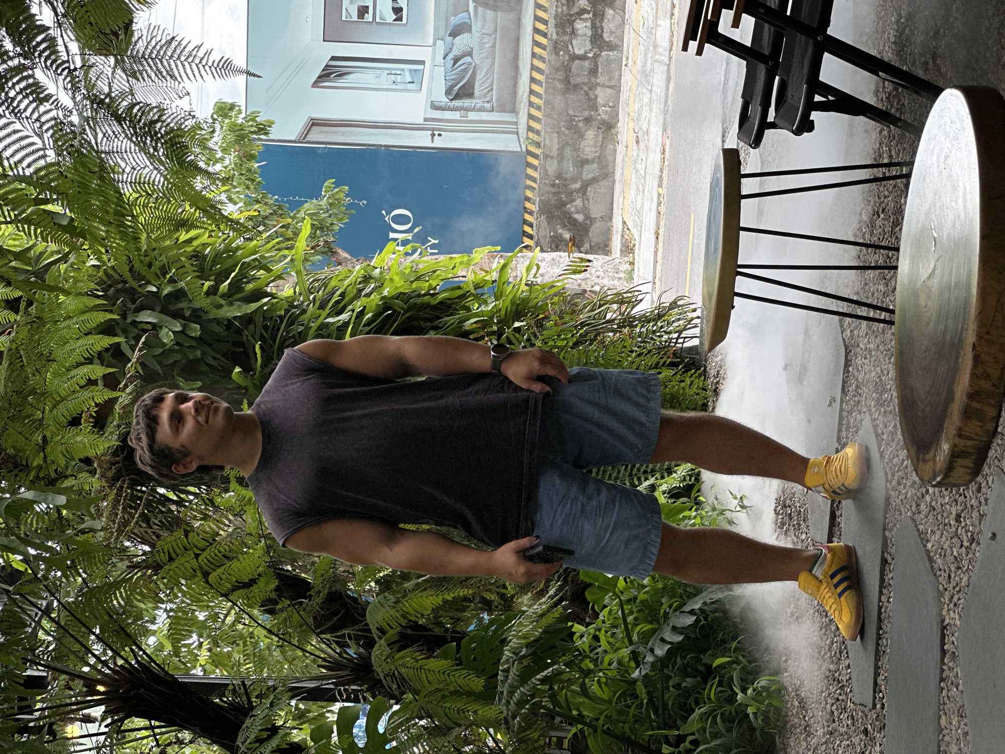 In Đà Nẵng, Vietnam, a young man stands in an outdoor seating area amidst lush greenery and a light mist. He is wearing a sleeveless dark gray t-shirt, light blue denim shorts, and bright yellow Adidas sneakers with blue stripes. He holds a dark-colored smartphone in his right hand and wears a watch on his left wrist. The background features a building with a large blue advertisement showcasing interior design, as well as a stone wall and sidewalk. Two black folding chairs and a small, light-colored table with thin black legs are visible in the foreground. The ground is covered with gravel and large, flat grey stepping stones.