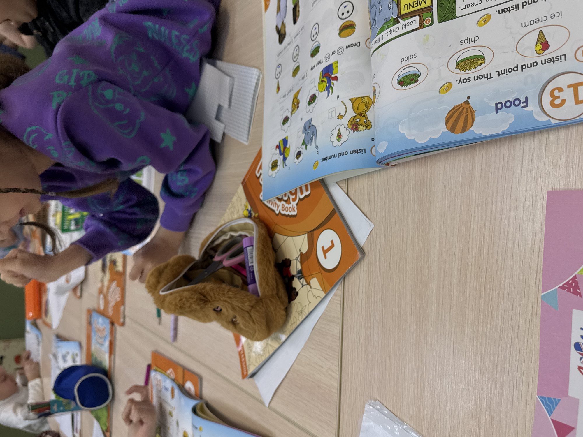 The scene appears to be a classroom setting in Perm, Russia, with children working on English lessons. There are children present at a table, with English activity books open in front of them. The books are titled "Activity Book" and feature images of food such as ice cream, chips, and salad, along with instructions like "Listen and point" and "Draw or Say." A fuzzy pencil case filled with school supplies sits on top of the books. A girl with braided hair wears a purple hoodie with green designs. Other details include pens, a blue pencil case, and possibly other school supplies scattered on the table.