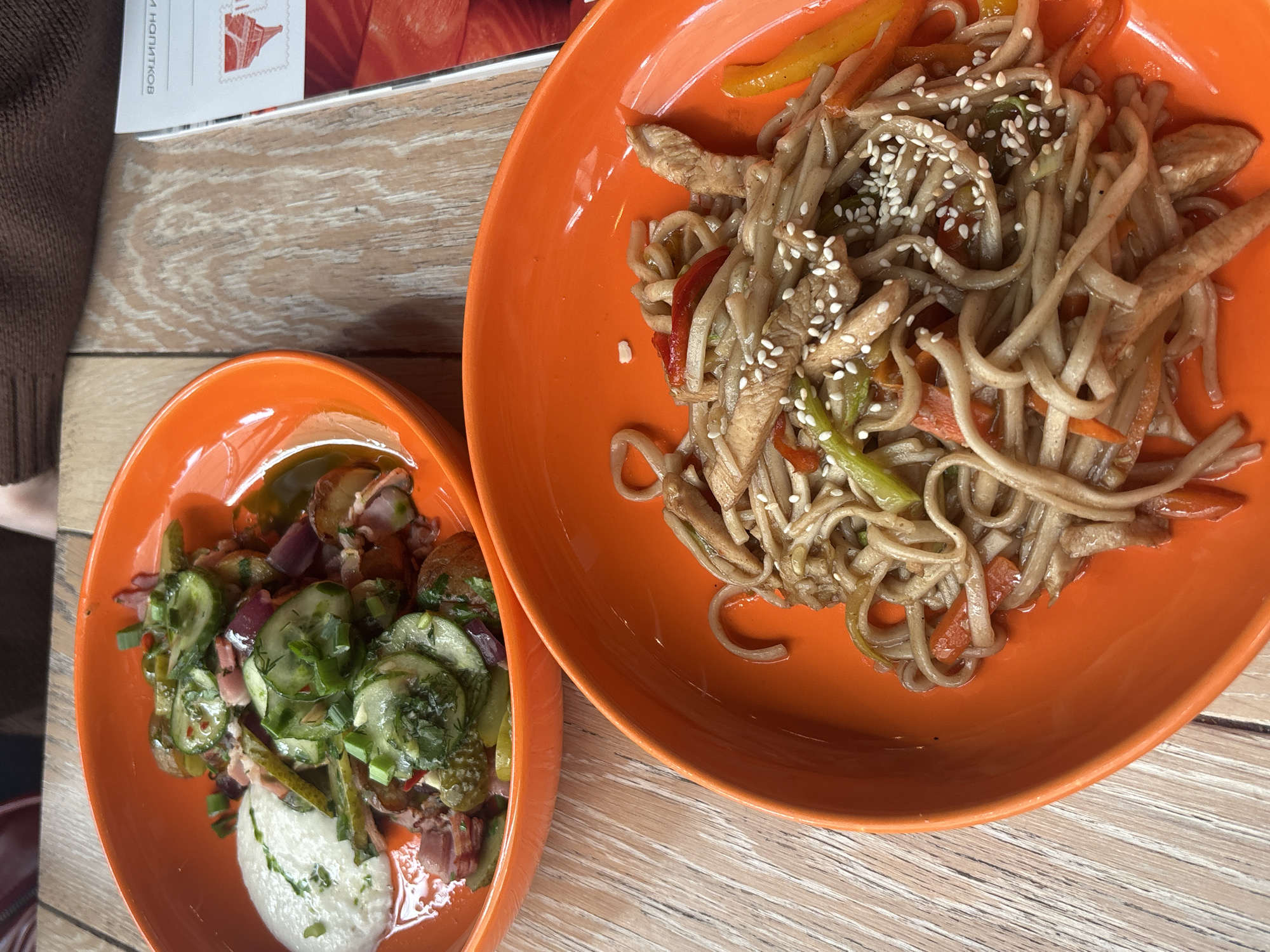 On a wooden table, two orange bowls present contrasting culinary arrangements. The larger bowl contains noodles stir-fried with what appears to be chicken and julienned vegetables, garnished with sesame seeds. The smaller bowl holds a salad of potatoes, sliced cucumbers, red onion, and herbs, likely dill and green onion, with a dollop of a white, creamy sauce. A portion of a menu or printed document rests partially visible in the upper left corner, along with a sleeve or garment of dark brown color. The setting suggests a restaurant or cafe environment. Location is Unknown, Russia.