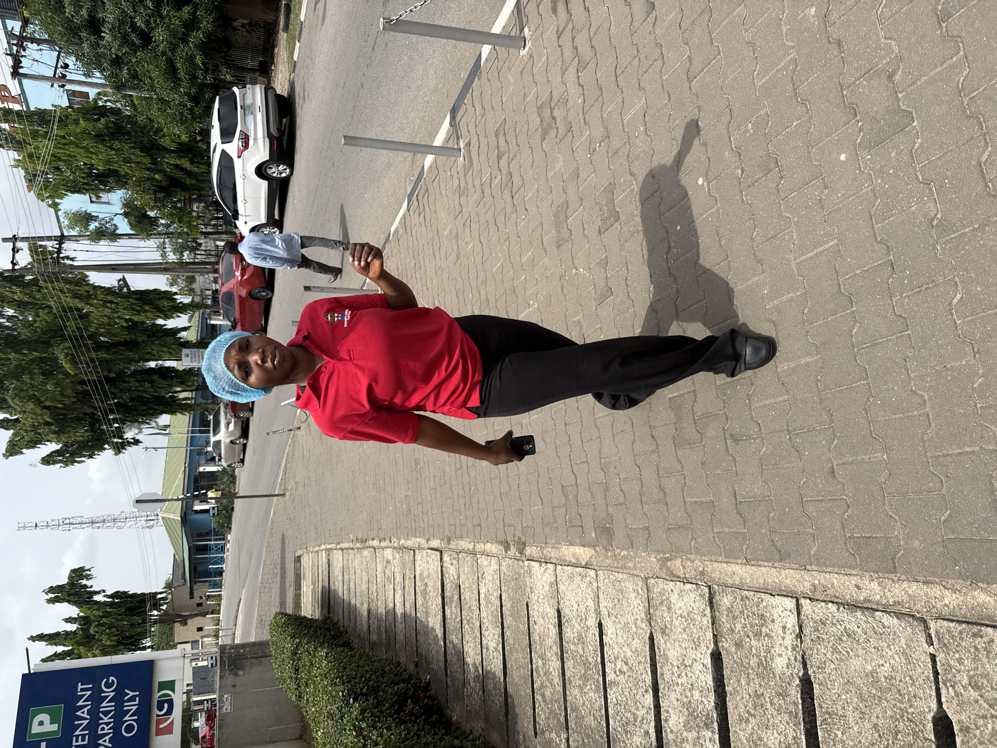 A woman in a red shirt, black pants, and a blue hairnet stands on a brick-paved sidewalk. She is holding a smartphone in her right hand. Behind her are parked cars, green trees, and a blue and white sign labeled "Tenant Parking Only." A man in a light blue shirt and dark pants is walking away in the background. The location is in Abuja, Nigeria. A communication tower and various buildings are visible in the distance.