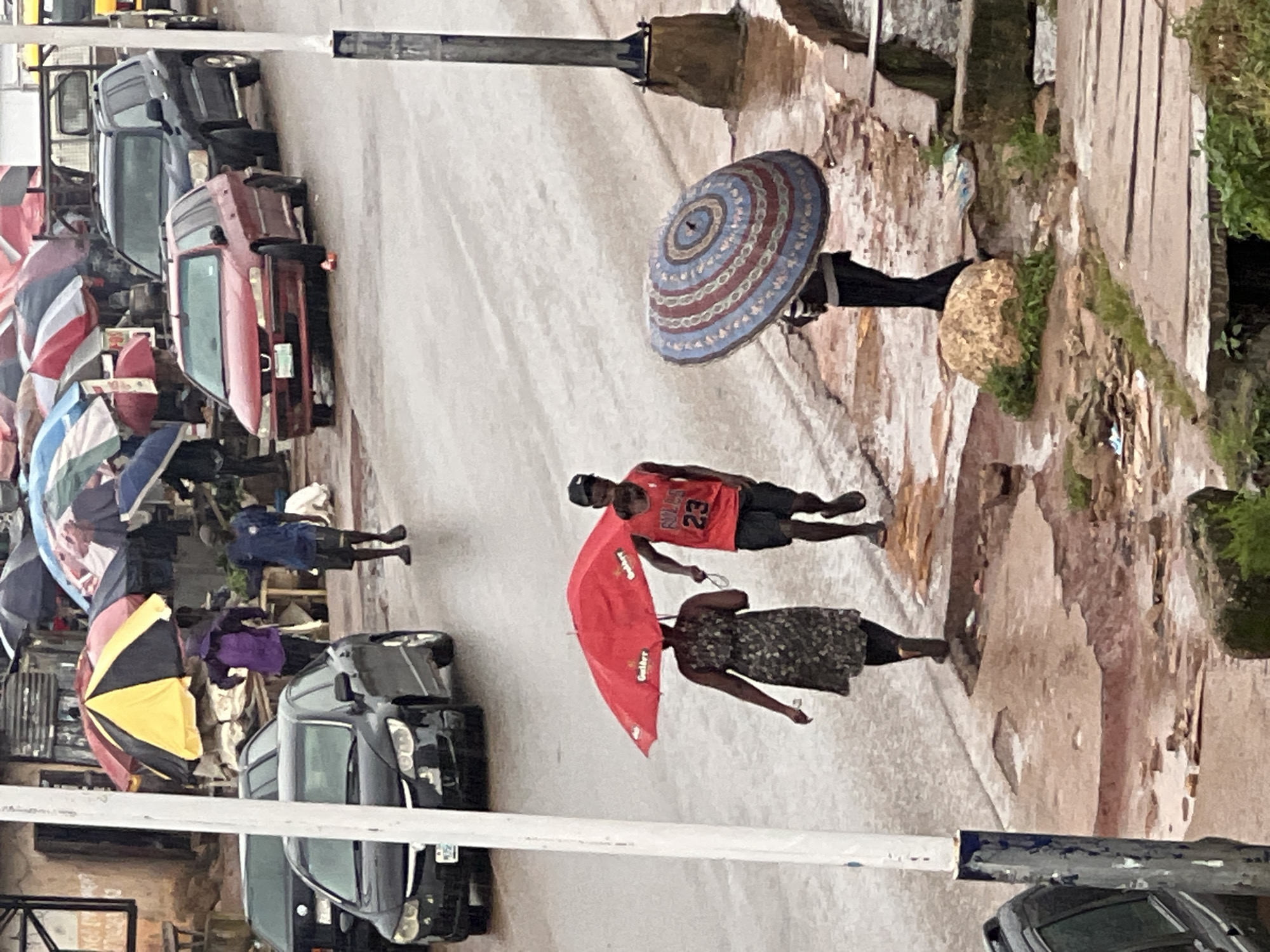 This image depicts a street scene in Jos, Nigeria during a heavy downpour. The wet asphalt road is visible, with puddles and reflections indicating the rain. Several vehicles are present, including a black sedan on the left, a red station wagon in the middle, and a black SUV parked behind it. In the background, market stalls with colorful umbrellas are set up, some of which are closed or tilted due to the rain.
In the foreground, three individuals are navigating the weather. A woman in a dark patterned dress and black leggings is walking under a bright red umbrella, holding its handle. Beside her, a man wearing a red tank top with the number "23" on it and black shorts is also walking. Further to the right, a person is holding a large, circular umbrella with a patterned design of concentric circles in red, blue, and white. The ground appears to be a mix of concrete and dirt, with some areas of exposed earth and scattered debris. The overall atmosphere is wet and overcast, characteristic of a rainy day in Jos.