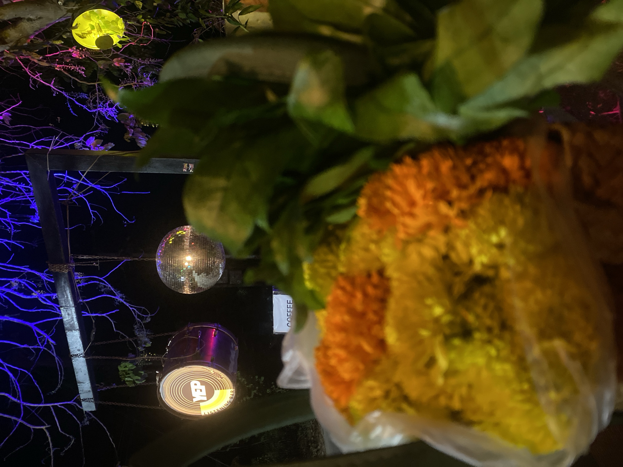 The image depicts an outdoor scene in Baan Don Sai, Thailand, at night. A partially obscured bouquet of yellow and orange flowers with green foliage is in the foreground. In the background, a metal frame structure is visible with branches illuminated by blue and pink lighting. Hanging from the frame are a disco ball and a sign that reads "YEP LISTENING BAR," backlit with yellow light. A sign reading "COFFEE" is barely visible behind the flowers. Above the metal structure is a yellow spherical light, further obscured by foliage.